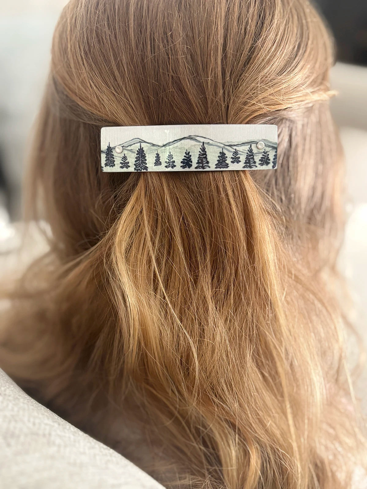 A close-up of a person's blonde hair secured with a rectangular hair clip featuring a black mountain and pine tree landscape design on a metallic background.