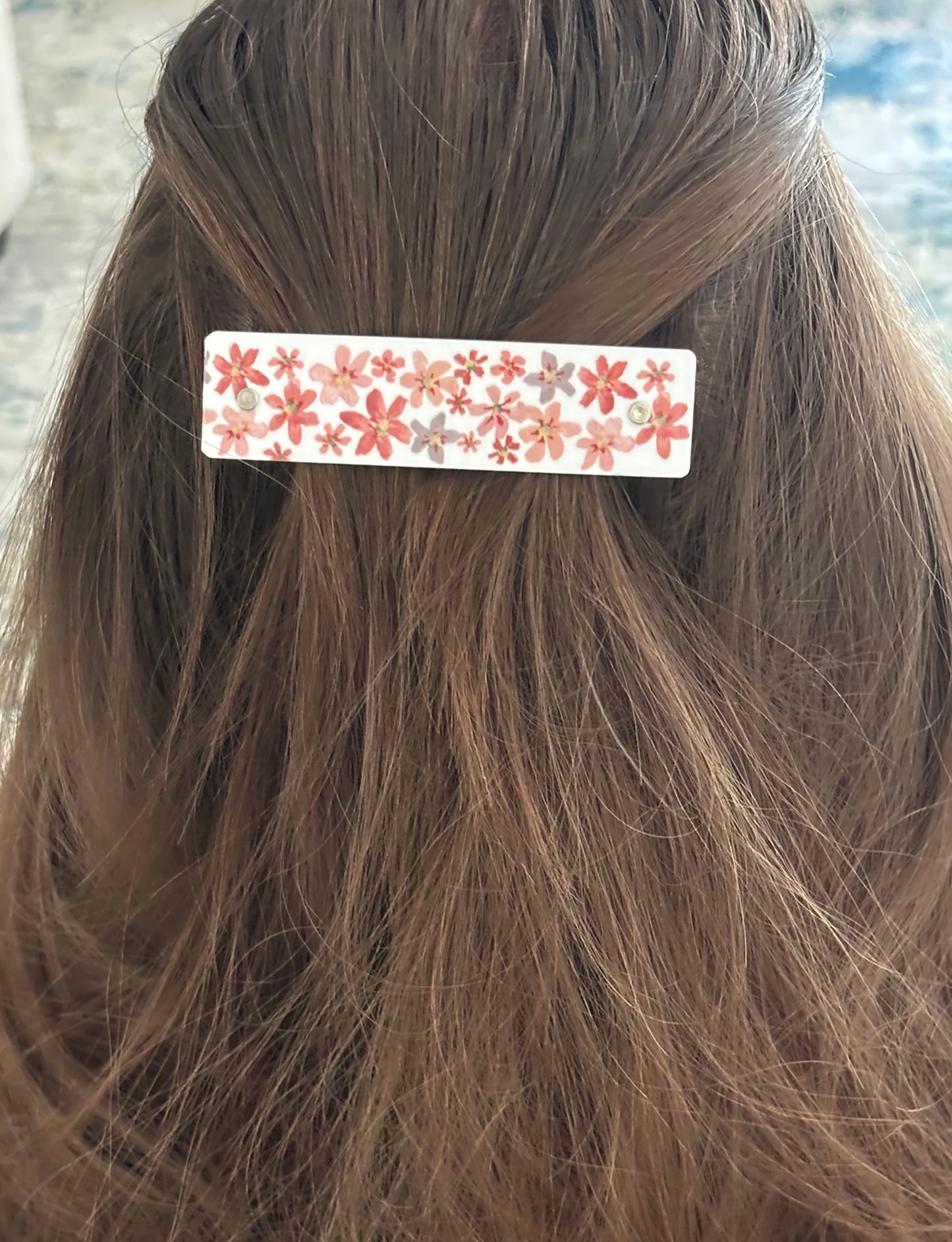 A close-up of a person's brown hair secured with a rectangular hair clip decorated with a pink, red, and orange floral pattern on a white background.