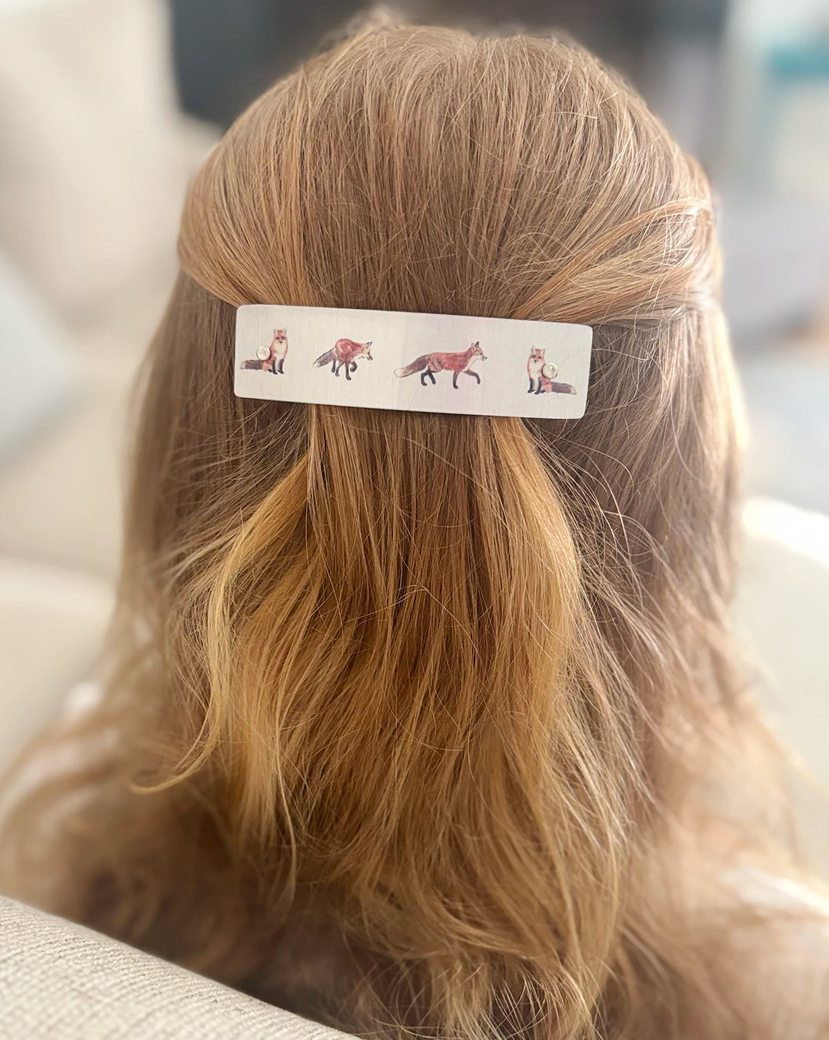A close-up of a person's blonde hair secured with a silver rectangular hair clip decorated with small illustrated foxes, shown from the back.