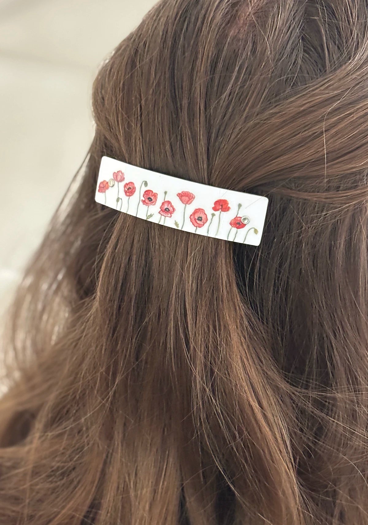 A close-up of a white boutique handmade hair clip decorated with a red poppy floral pattern, holding back a section of medium brown hair.