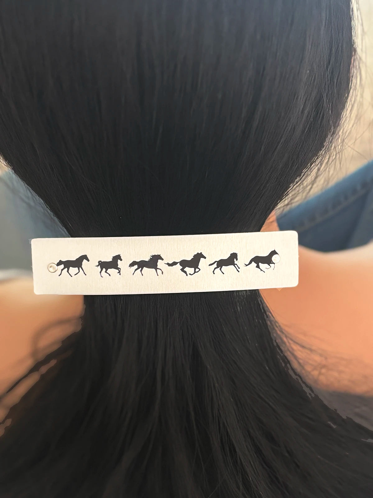 A close-up of a person's dark hair secured with a silver rectangular hair clip featuring a row of black galloping horse silhouettes.