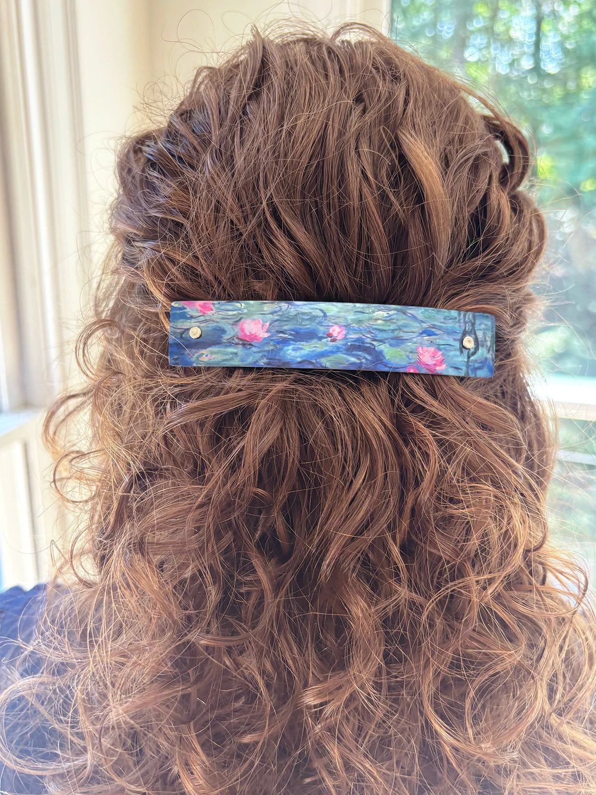 A close-up back view of a person with long, wavy light brown hair wearing a rectangular hair clip. The clip features a painted water lily design with pink flowers and blue-green water in a pond, secured with two small silver metal rivets.