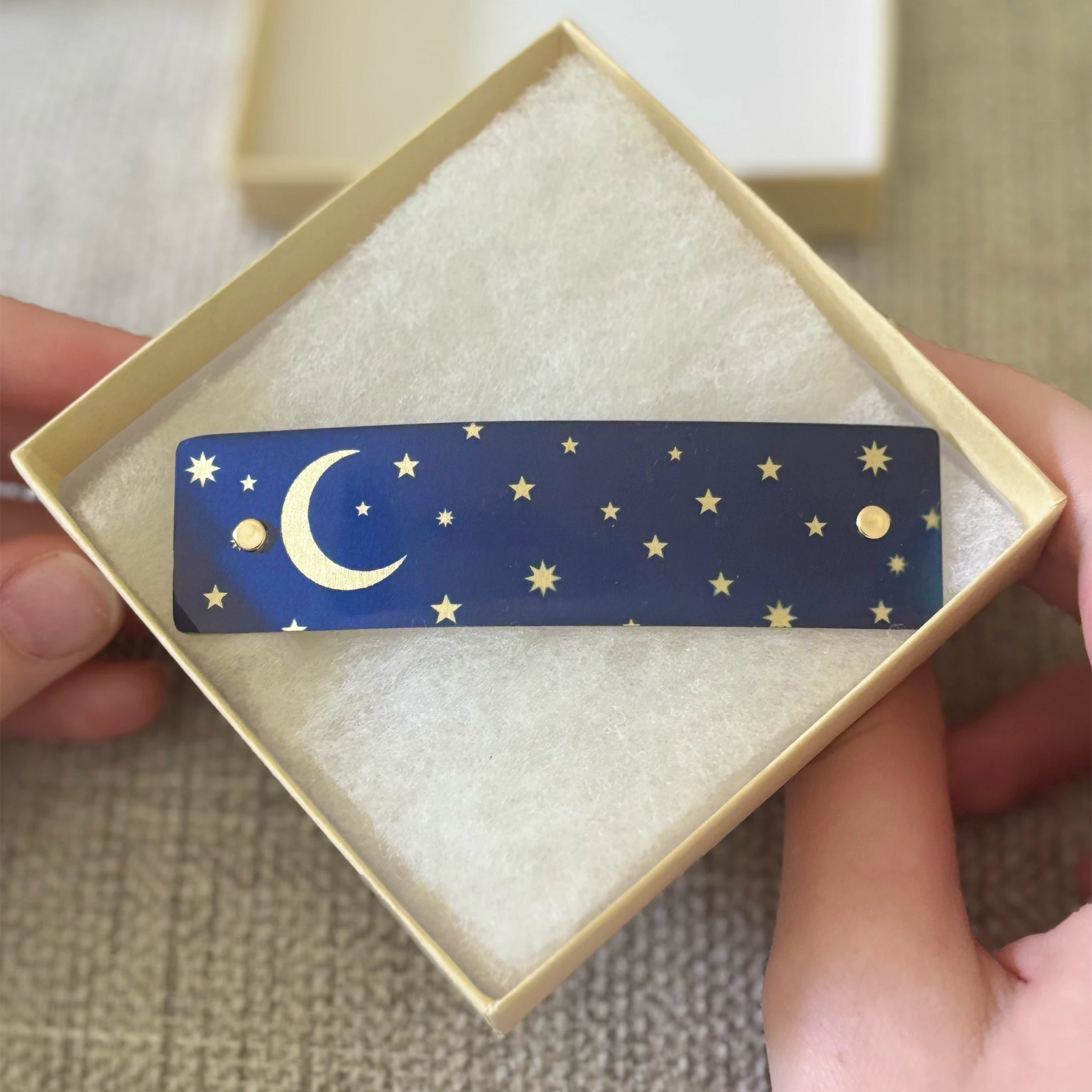 A rectangular navy blue hair barrette with a gold crescent moon and stars, presented in a cream-colored gift box held by hands.