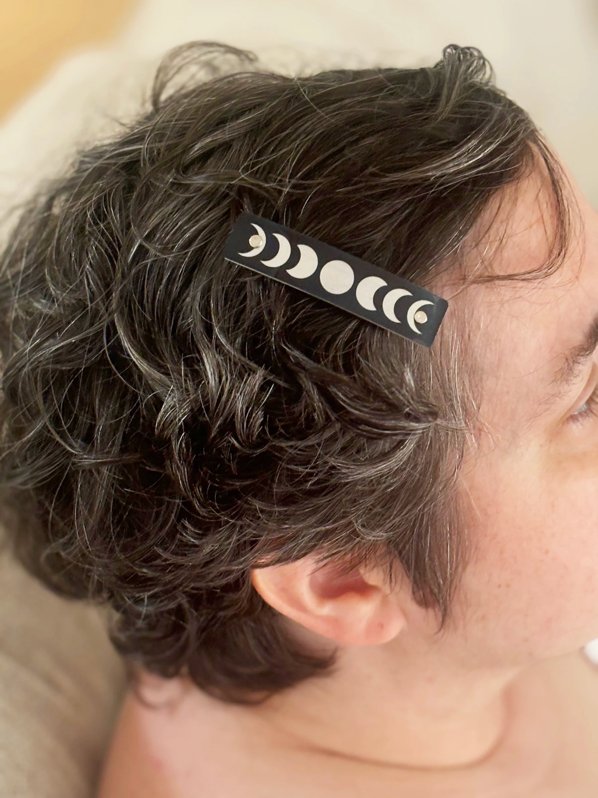 A close-up of a person's short, curly dark hair wearing a black hair clip decorated with a silver moon phase pattern.