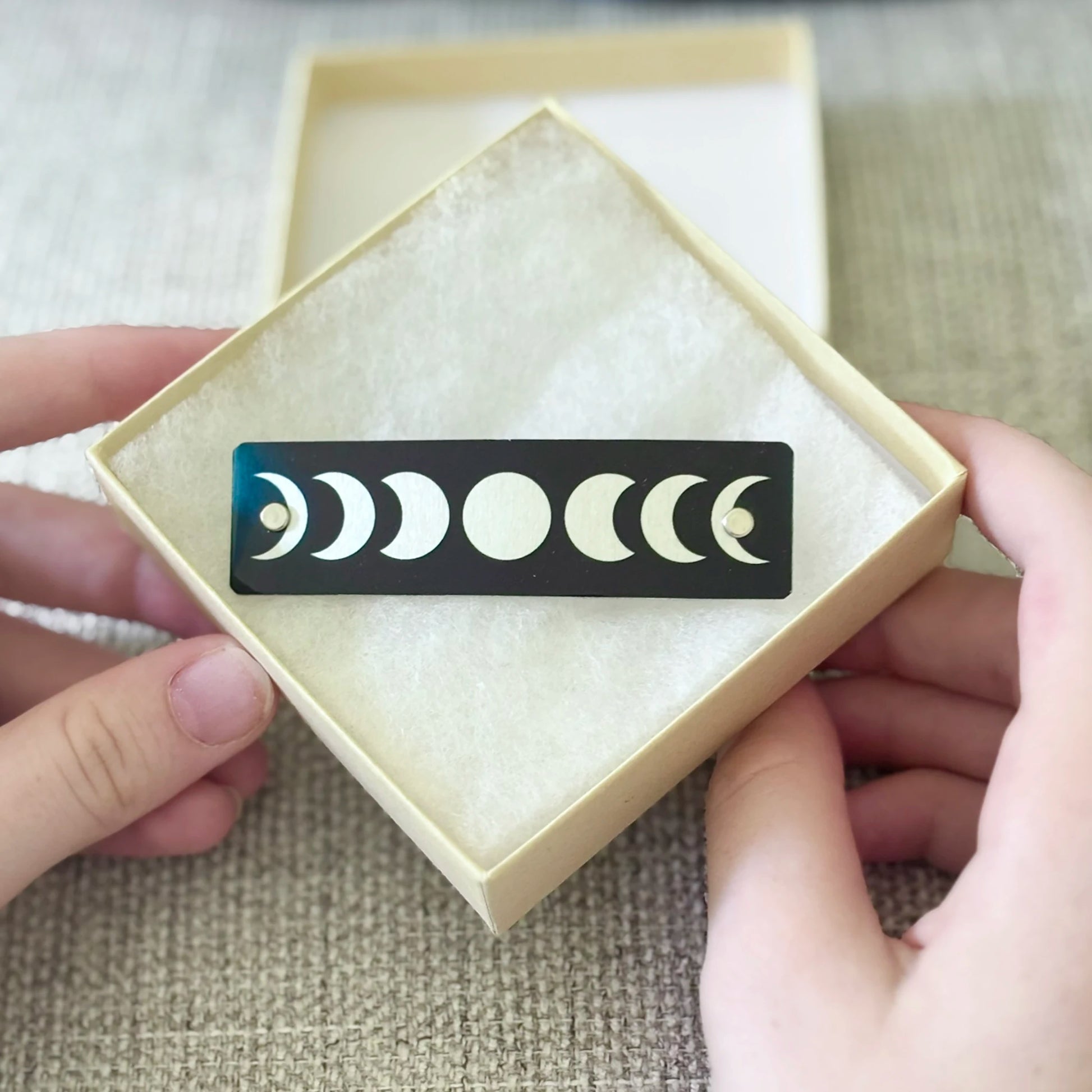 A black rectangular hair clip featuring a moon phase progression, held in a beige presentation box with two hands visible against a textured neutral background.