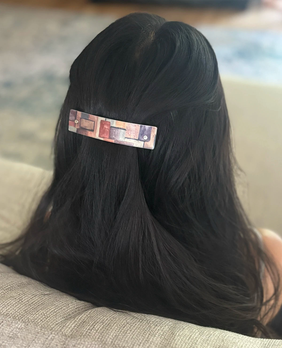 A close-up back view of a person with long, straight black hair wearing a slim rectangular hair clip with a multicolor geometric pattern.