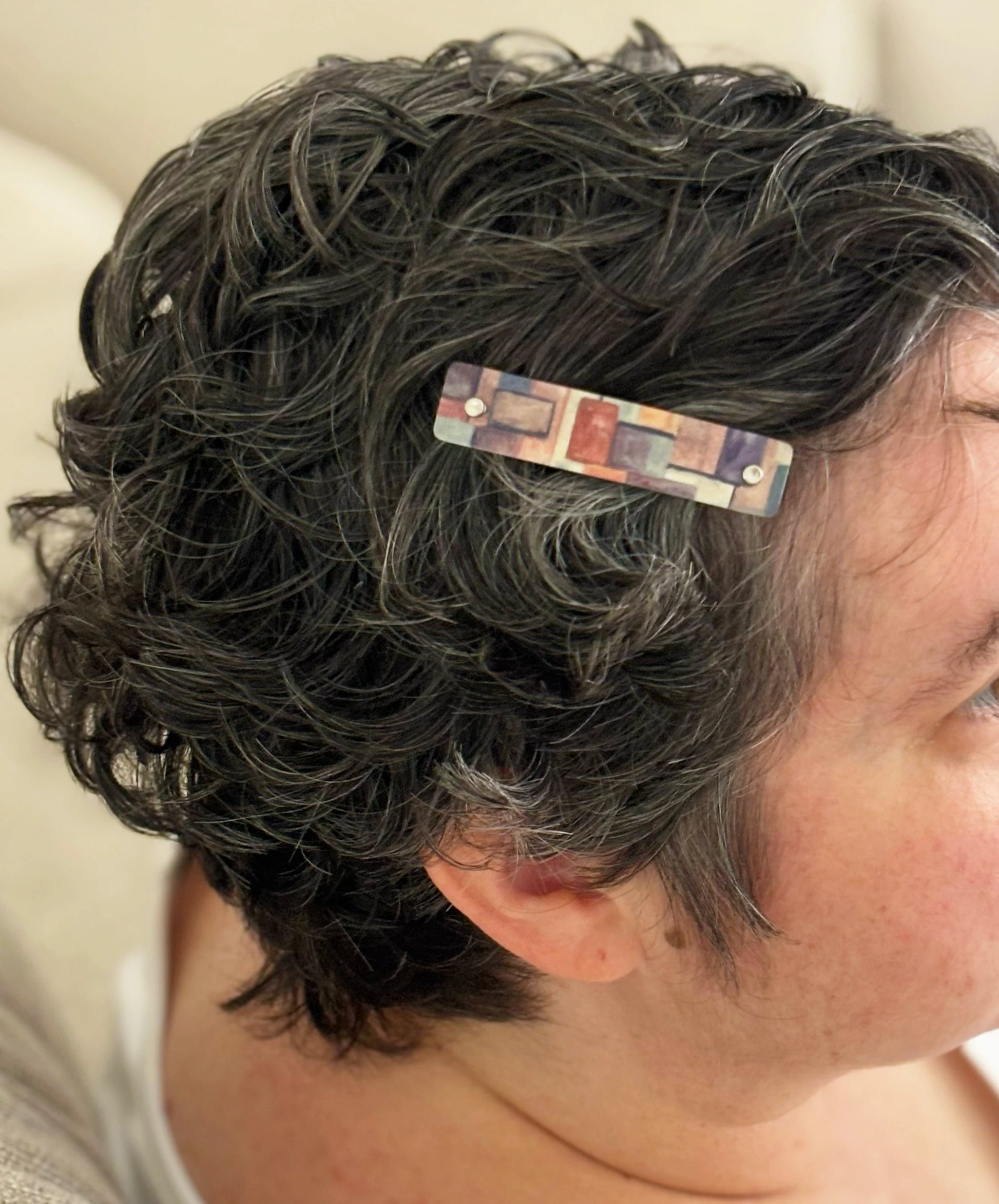 A close-up side profile of a person with short, curly dark gray hair wearing a rectangular hair clip with a multicolor geometric pattern.