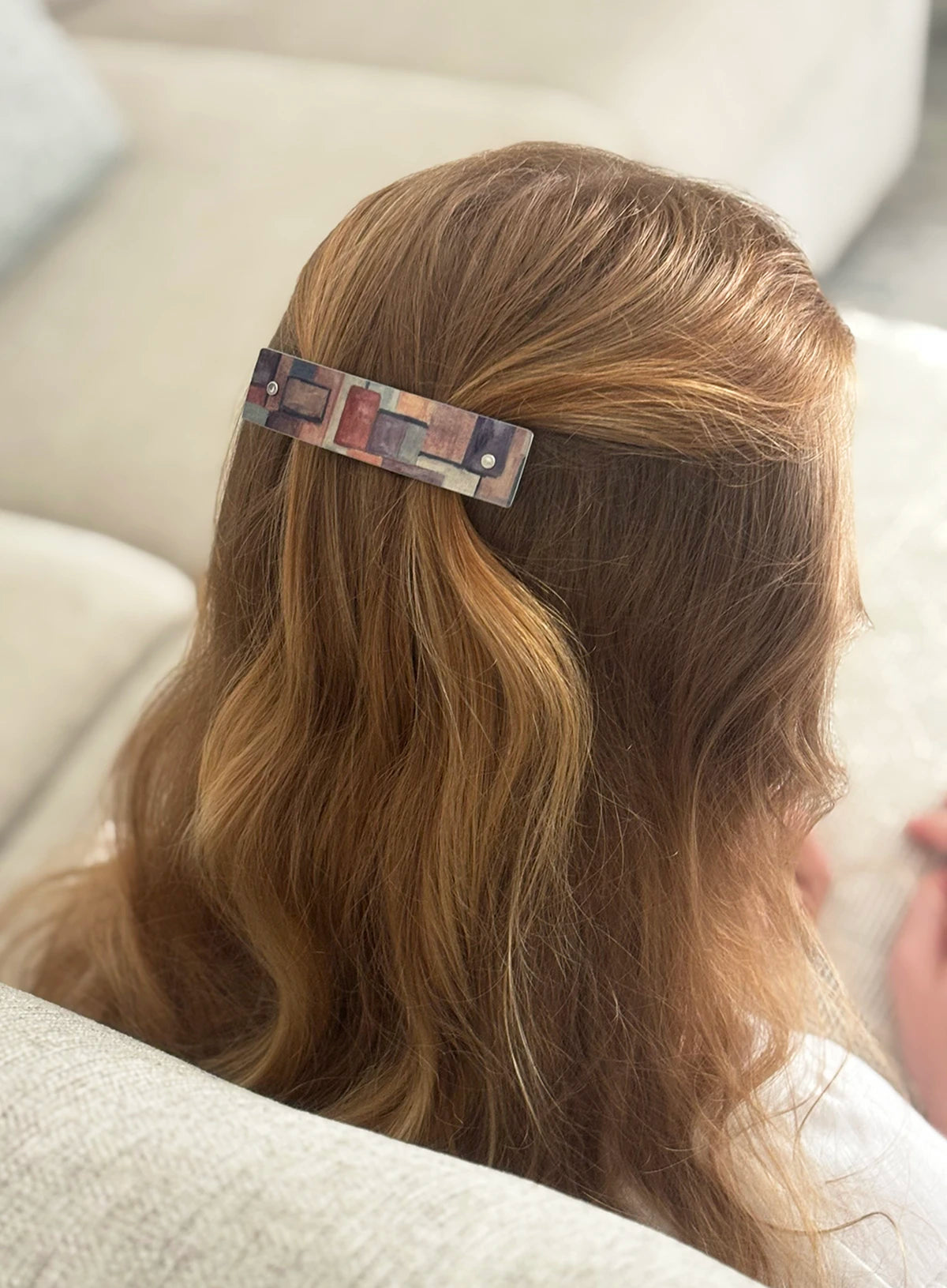 A close-up lifestyle shot of a person with long, wavy light brown hair wearing a rectangular hair clip. The clip features a multicolor abstract art print with soft earthy tones, and a small silver rivet in each end.