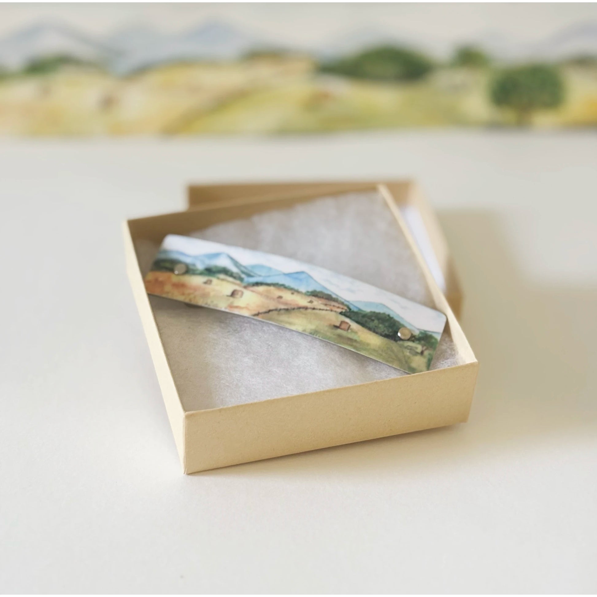 A small rectangular landscape art hair barrette with a watercolor-style scene of rolling hills and trees, resting inside an open beige gift box lined with white  cotton on a light surface.