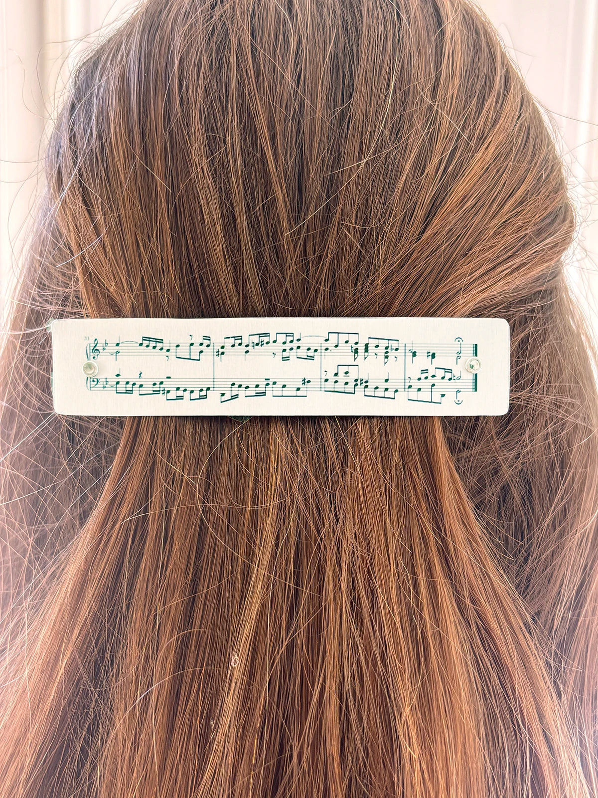 A close-up of a person's brown hair with a silver rectangular hair clip featuring printed musical notes and staff lines.