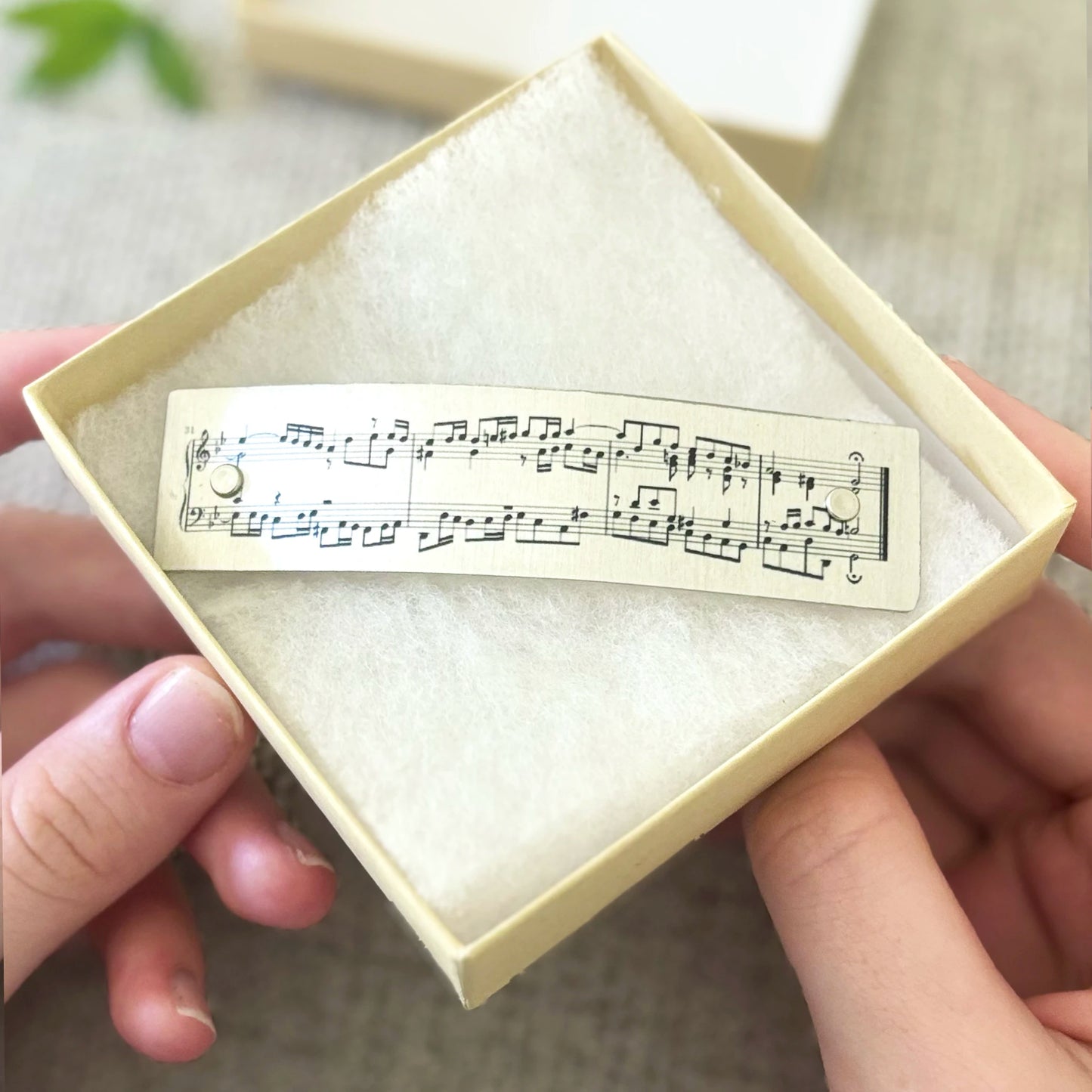 A person holds an open cream-colored jewelry box containing a French barrette hair clip printed with classical music notation. The background is a softly blurred neutral indoor setting.