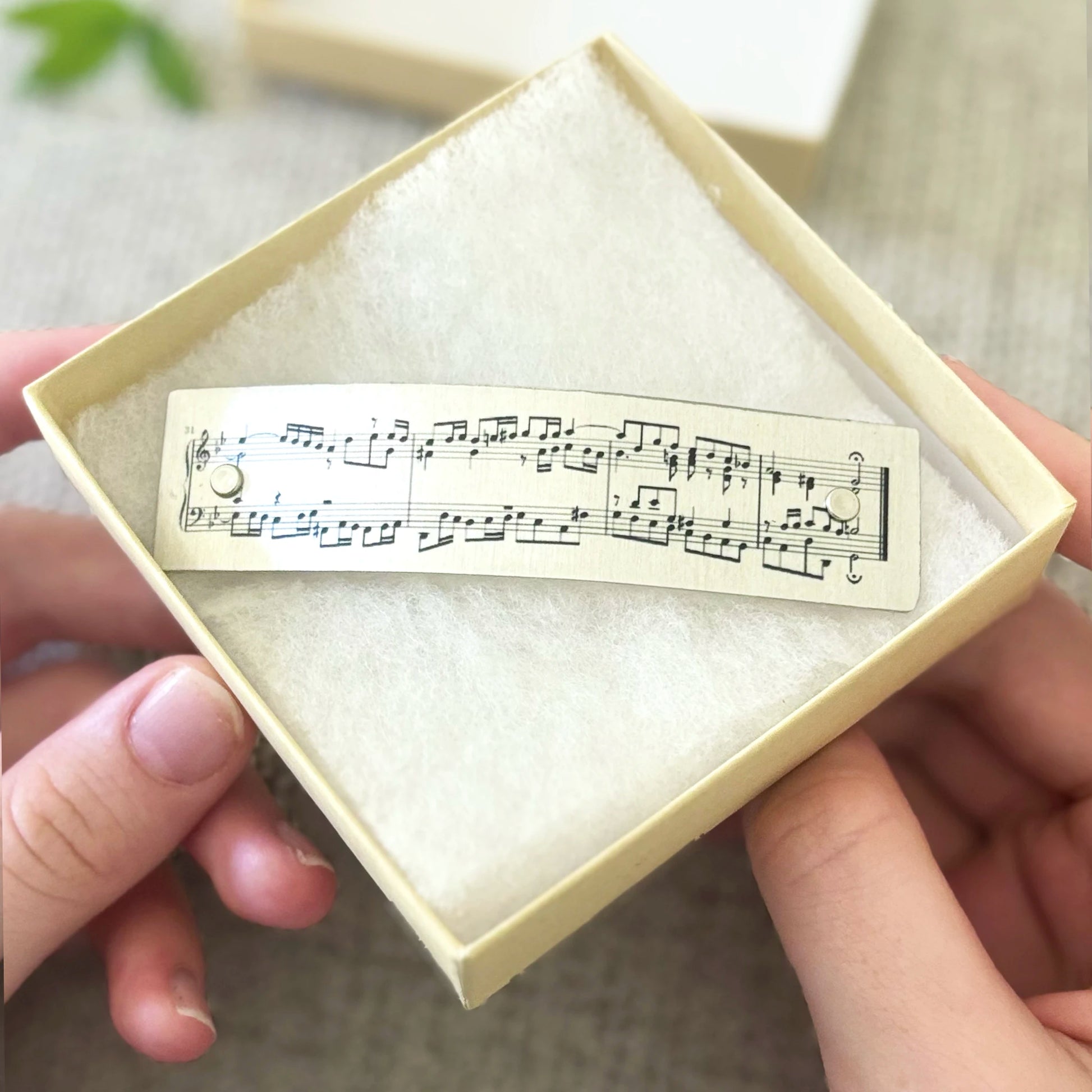 A person holds an open cream-colored jewelry box containing a French barrette hair clip printed with classical music notation. The background is a softly blurred neutral indoor setting.