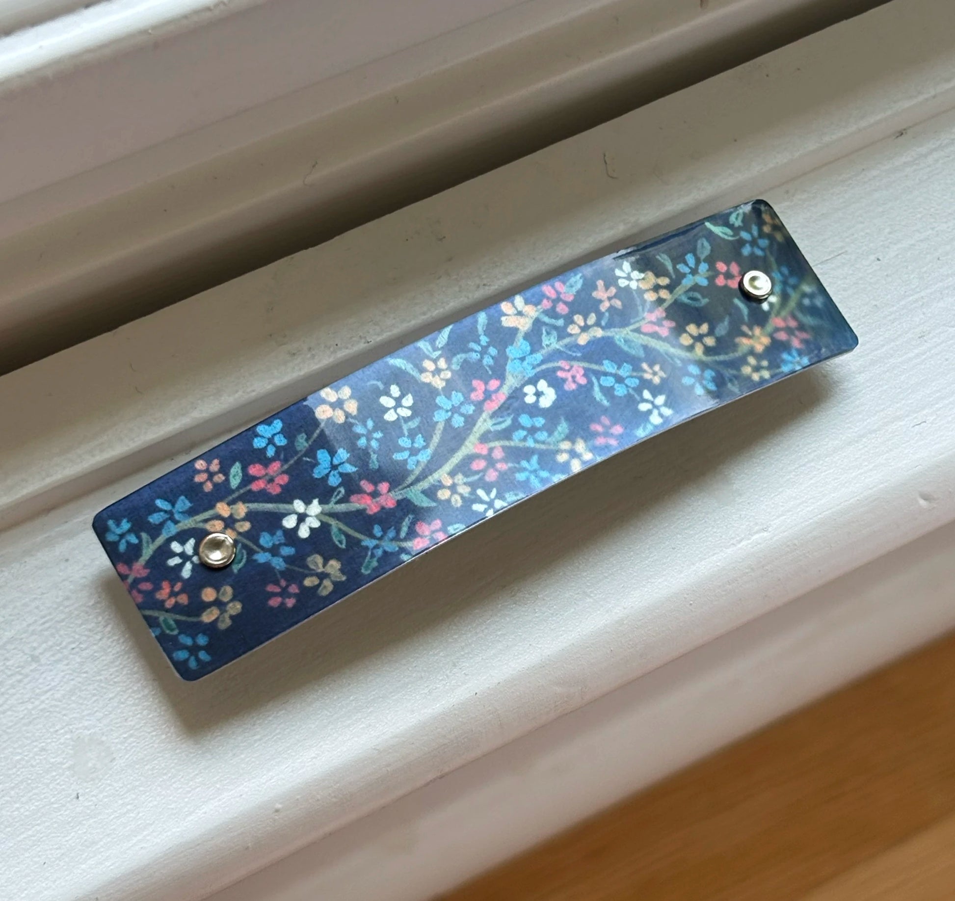 A long, narrow decorative bar hair clip with a blue floral pattern and two metal rivets, resting on a white wooden surface.