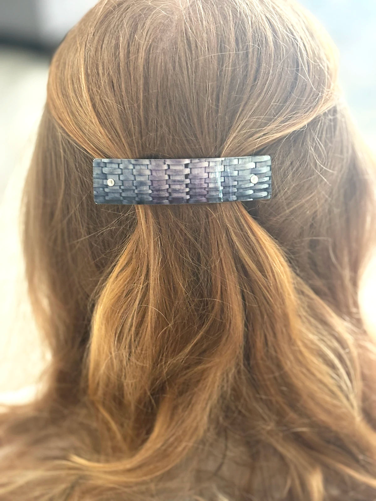 A close-up back view of a person's hair styled in a half-up ponytail, secured with a rectangular woven hair clip in a gray and silver pattern.