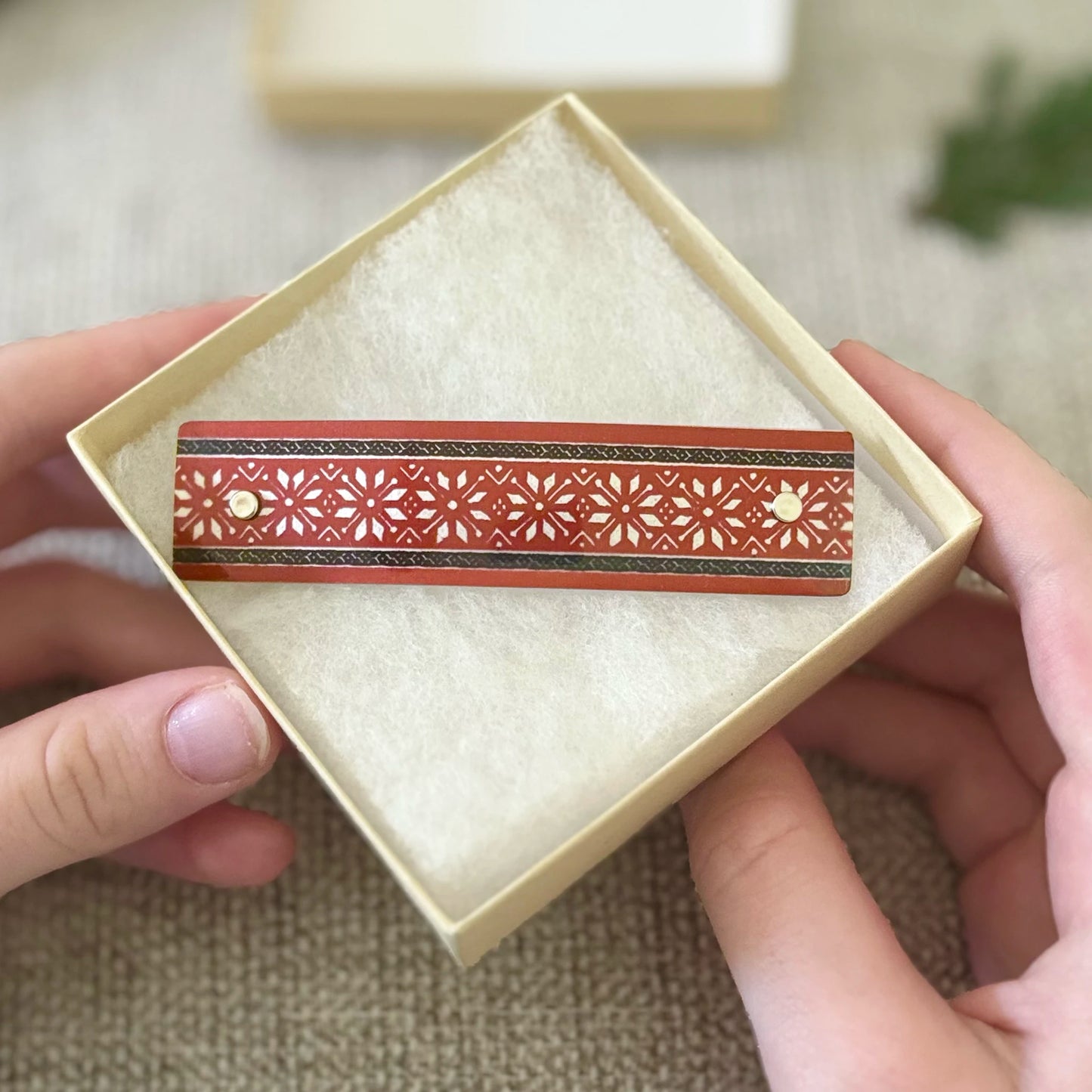A close-up of a hand holding an open cream-colored presentation box containing a decorative rectangular red Scandinavian fair isle pattern design with intricate star patterns and silver accents.