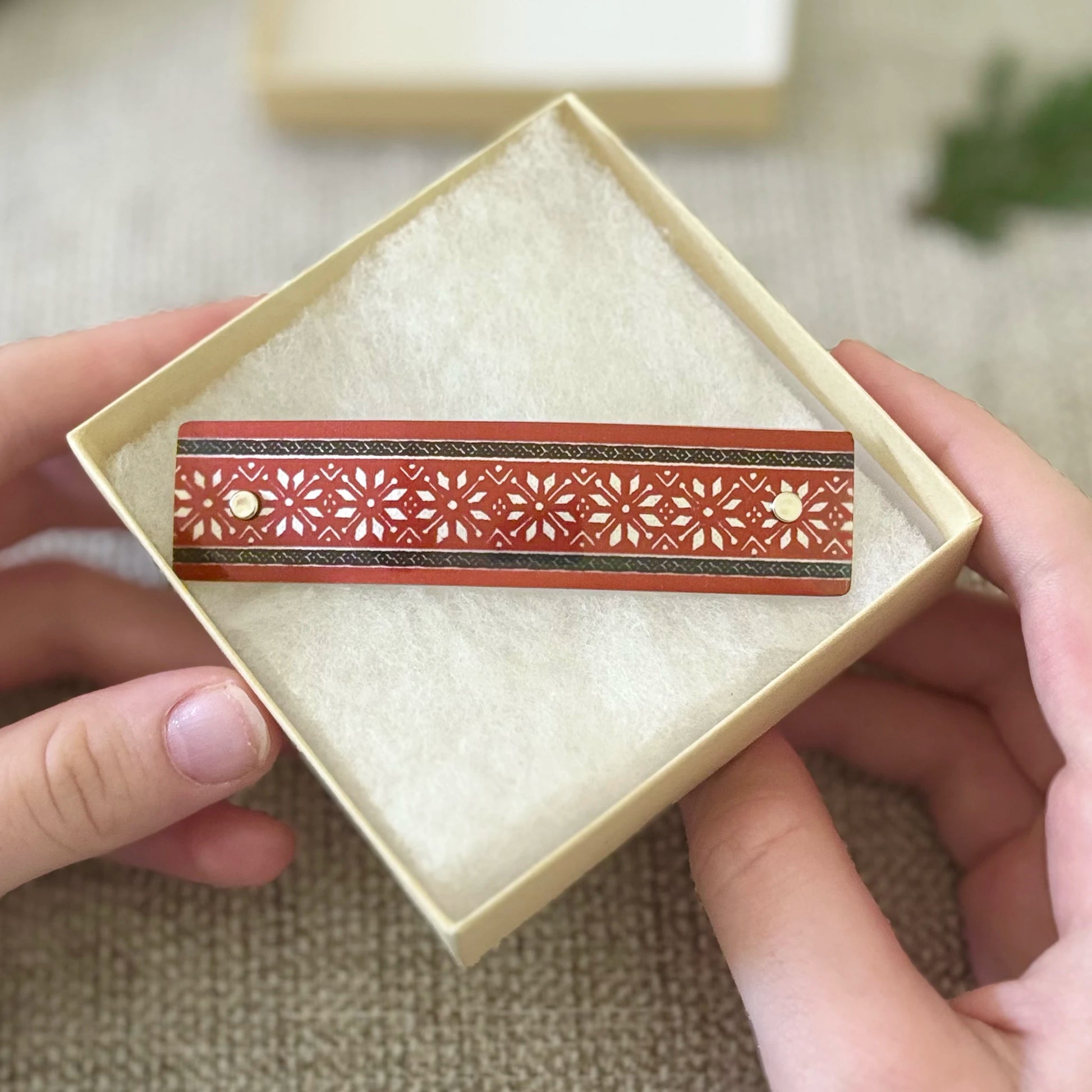 A close-up of a hand holding an open cream-colored presentation box containing a decorative rectangular red Scandinavian fair isle pattern design with intricate star patterns and silver accents.