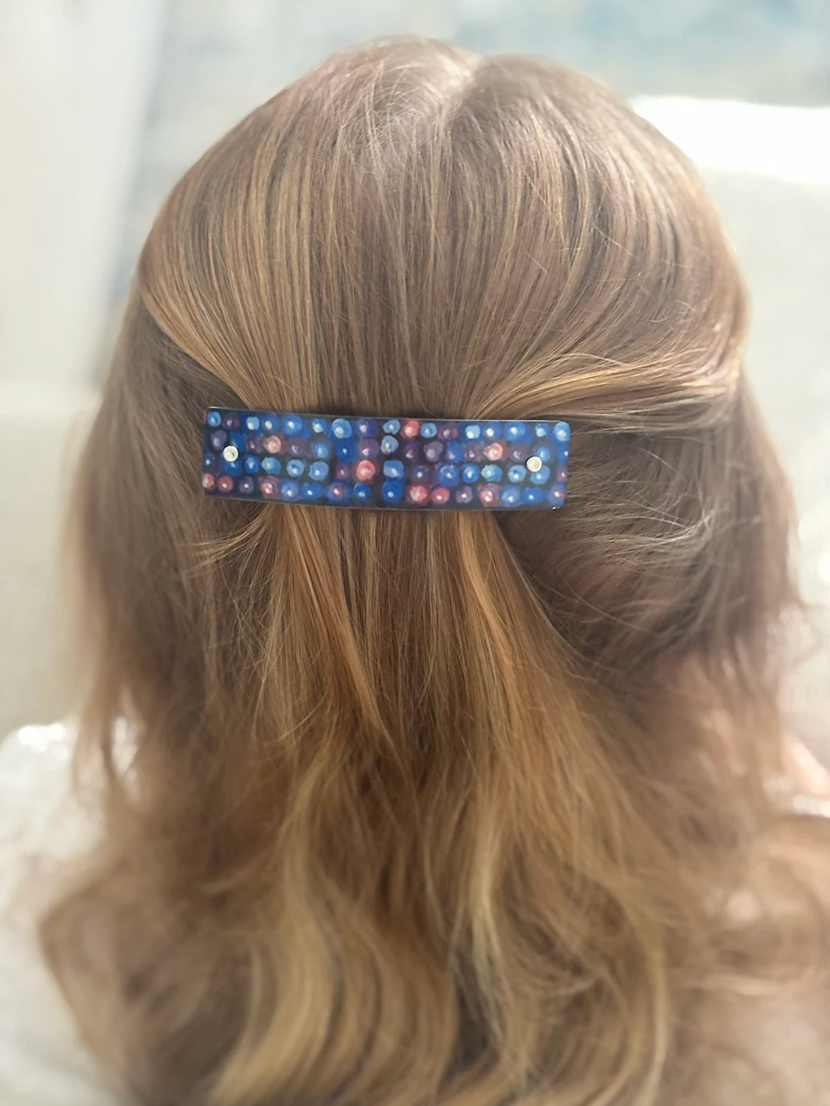 A close-up back view of a person with long light brown hair, wearing a small rectangular hair clip adorned with a blackberry pattern.
