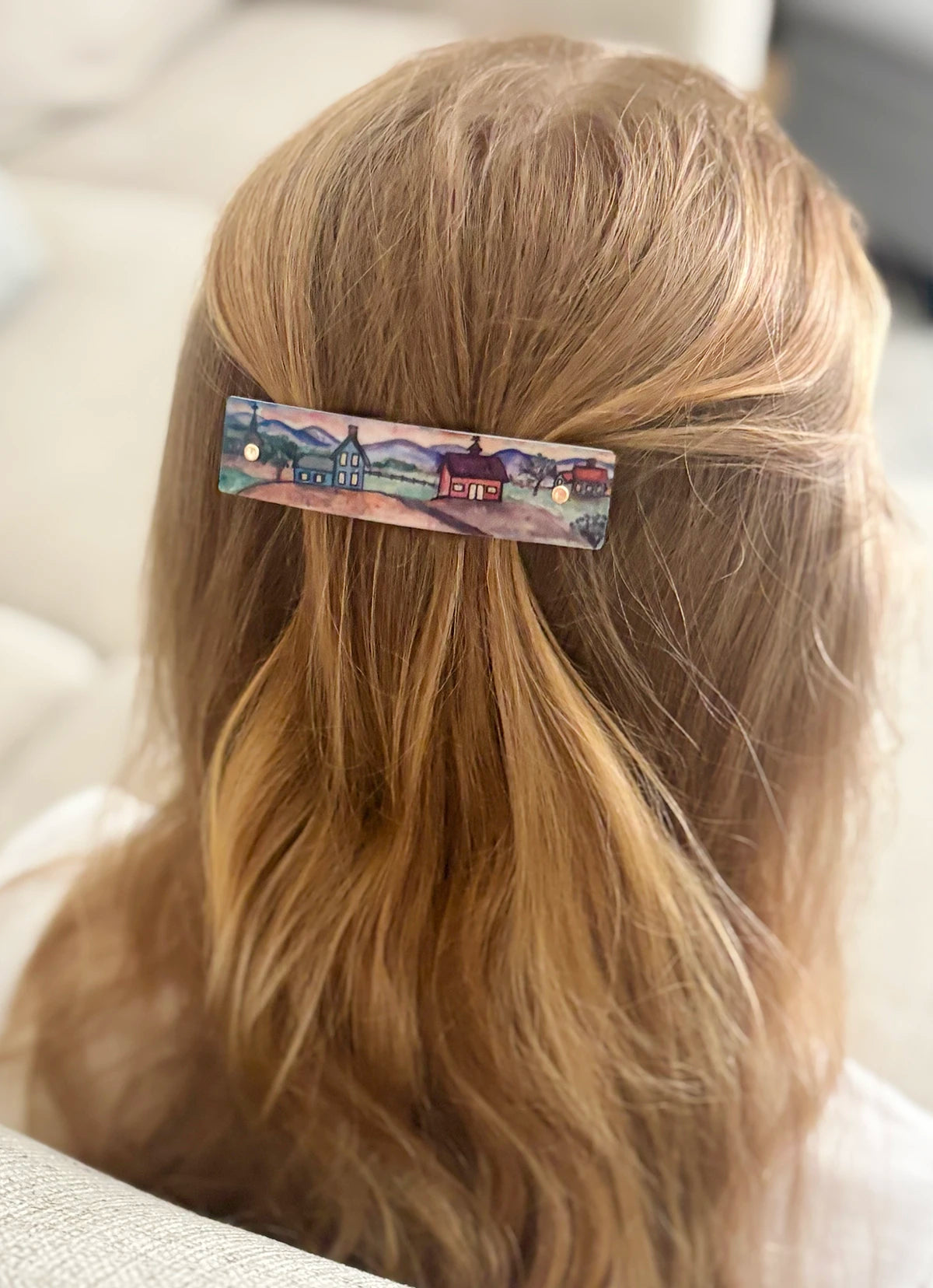 A blonde person shown from the back wearing a rectangular hair clip with a colorful painted village scene on it.