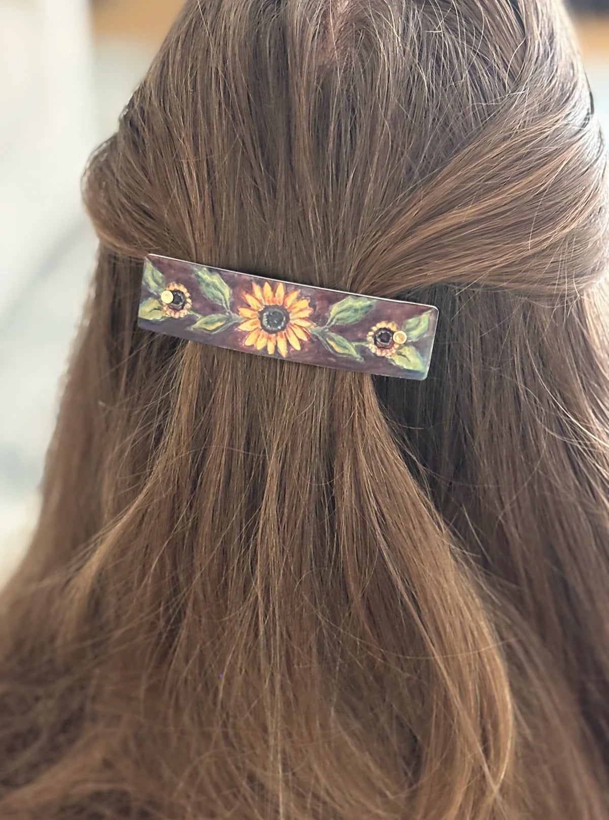 A close-up of a person's brown hair secured with a rectangular hair clip decorated with painted sunflowers and green leaves.