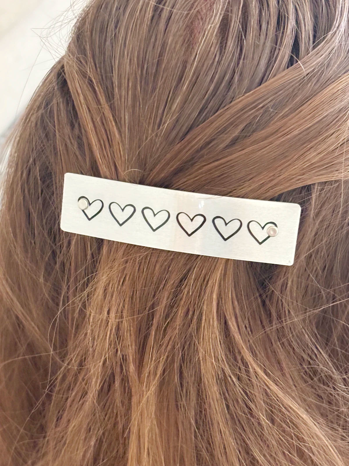 A close-up of light brown hair secured with a silver rectangular hair clip decorated with five small black heart outlines.