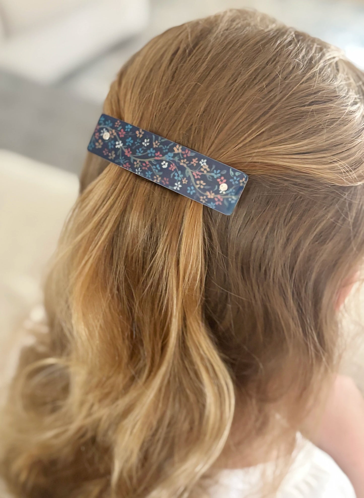 A close-up lifestyle shot of  blonde hair secured with a decorative blue barrette hair clip featuring a floral pattern.