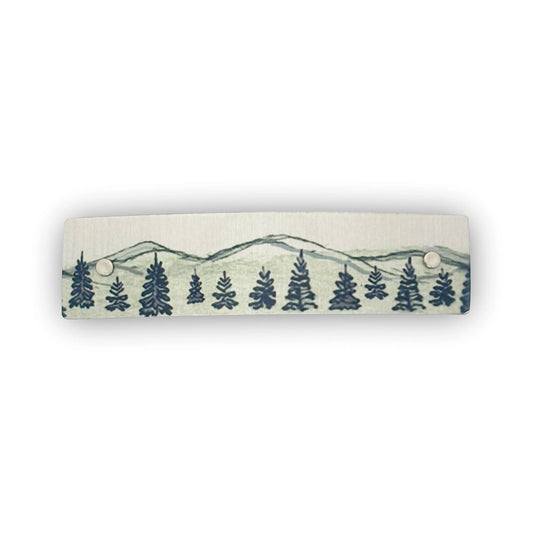 A silvery aluminum hair clip with a mountain and pine tree landscape shown on a white background.