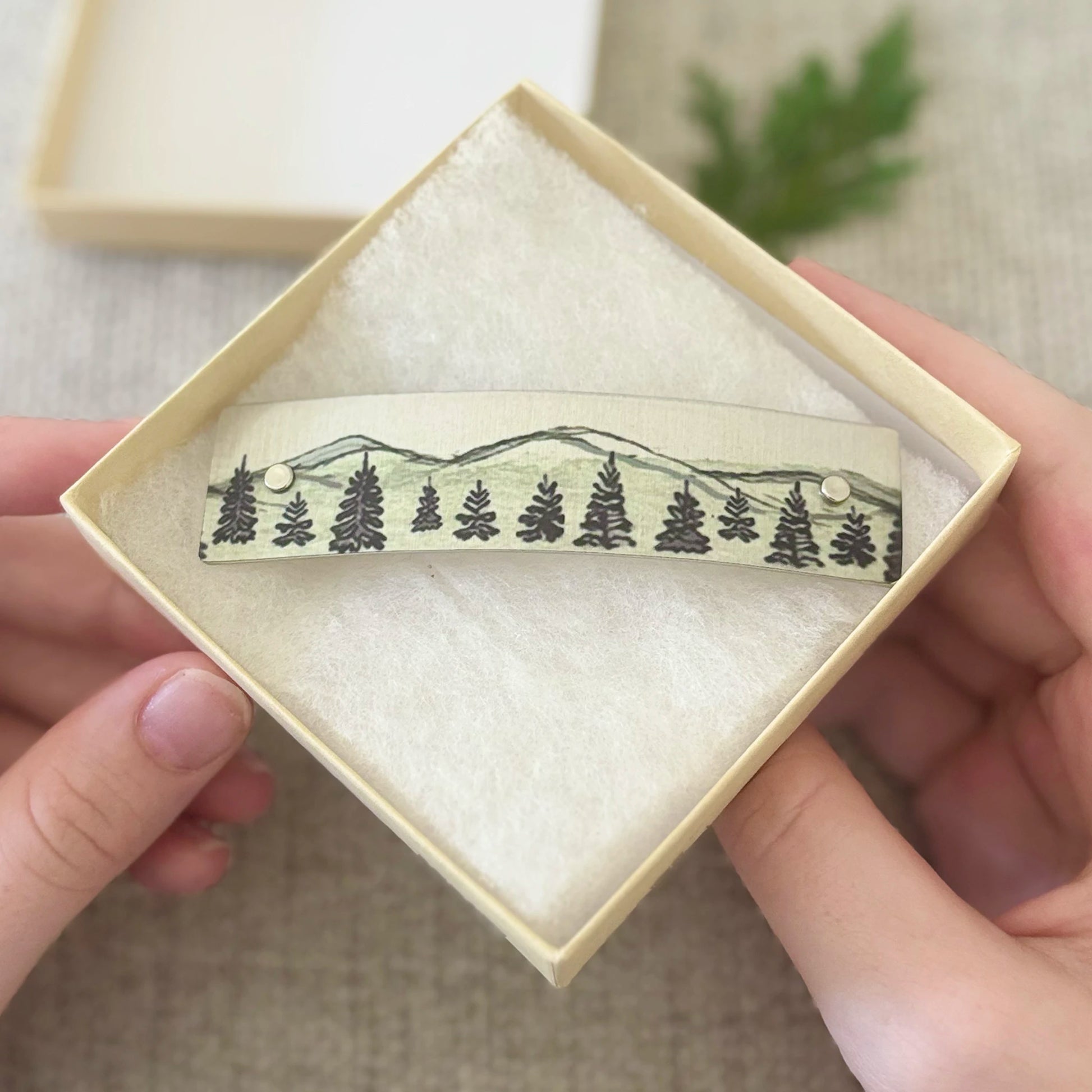 A close-up of  hands holding a small square jewelry box containing a French barrette hair clip decorated with a soft watercolor drawing of pine trees and mountains. The background is a soft beige fabric with a blurred green plant.