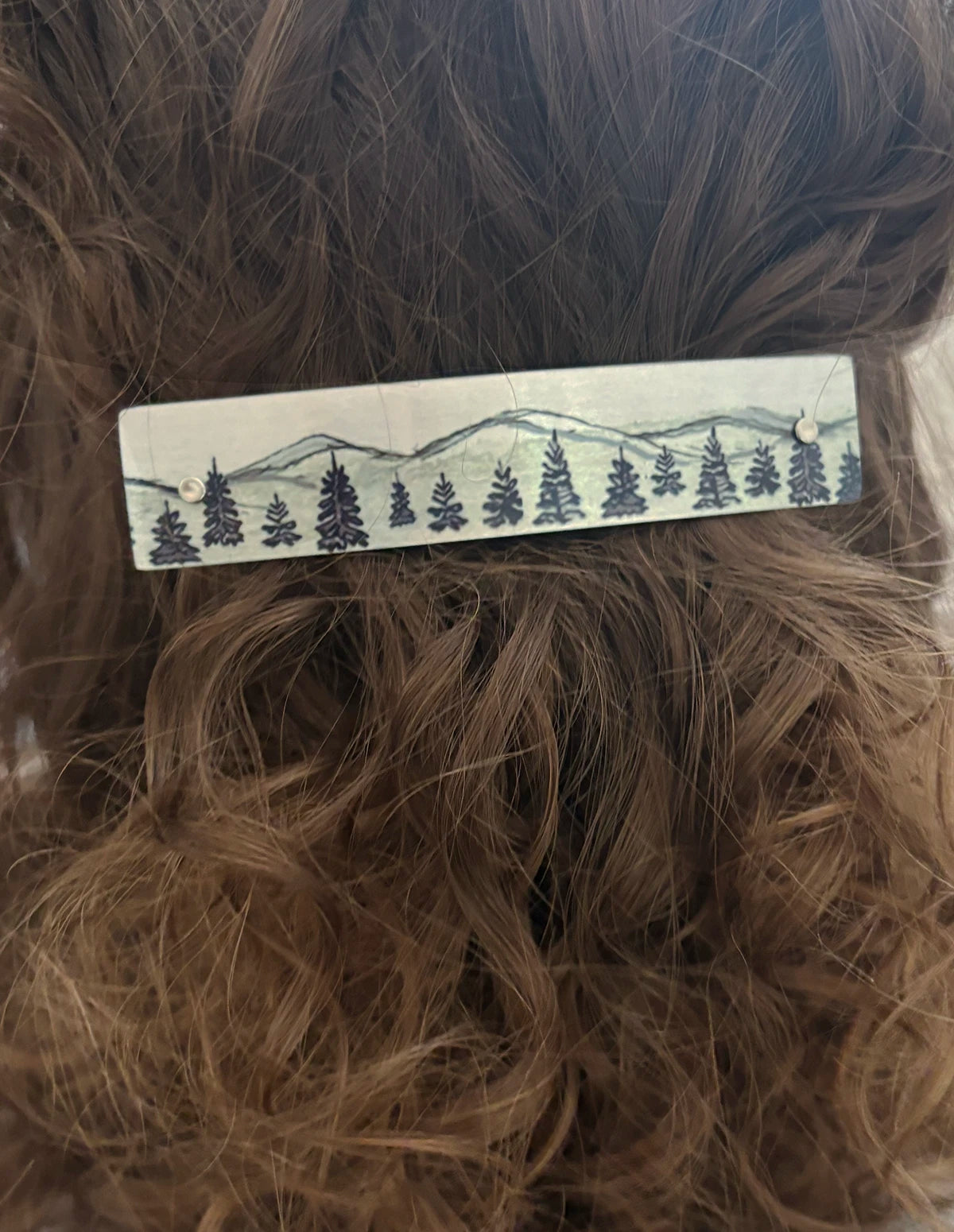 A close-up of brown hair with a rectangular metal hair clip featuring a printed winter mountain and pine tree design.