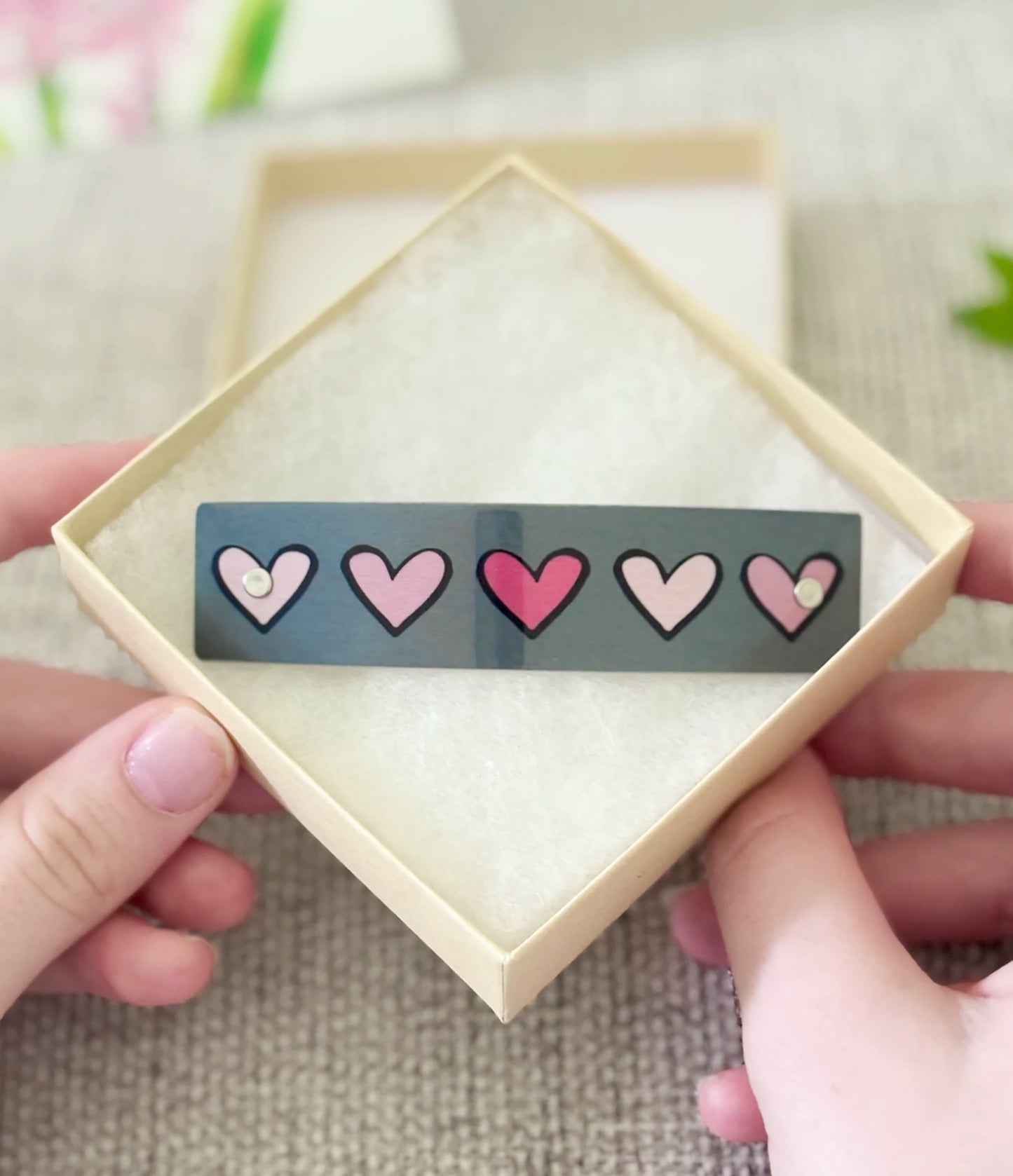 A close-up of a person's hands holding a square cream-colored gift box, displaying a row of pink hearts  with five pink hearts outlined in black.