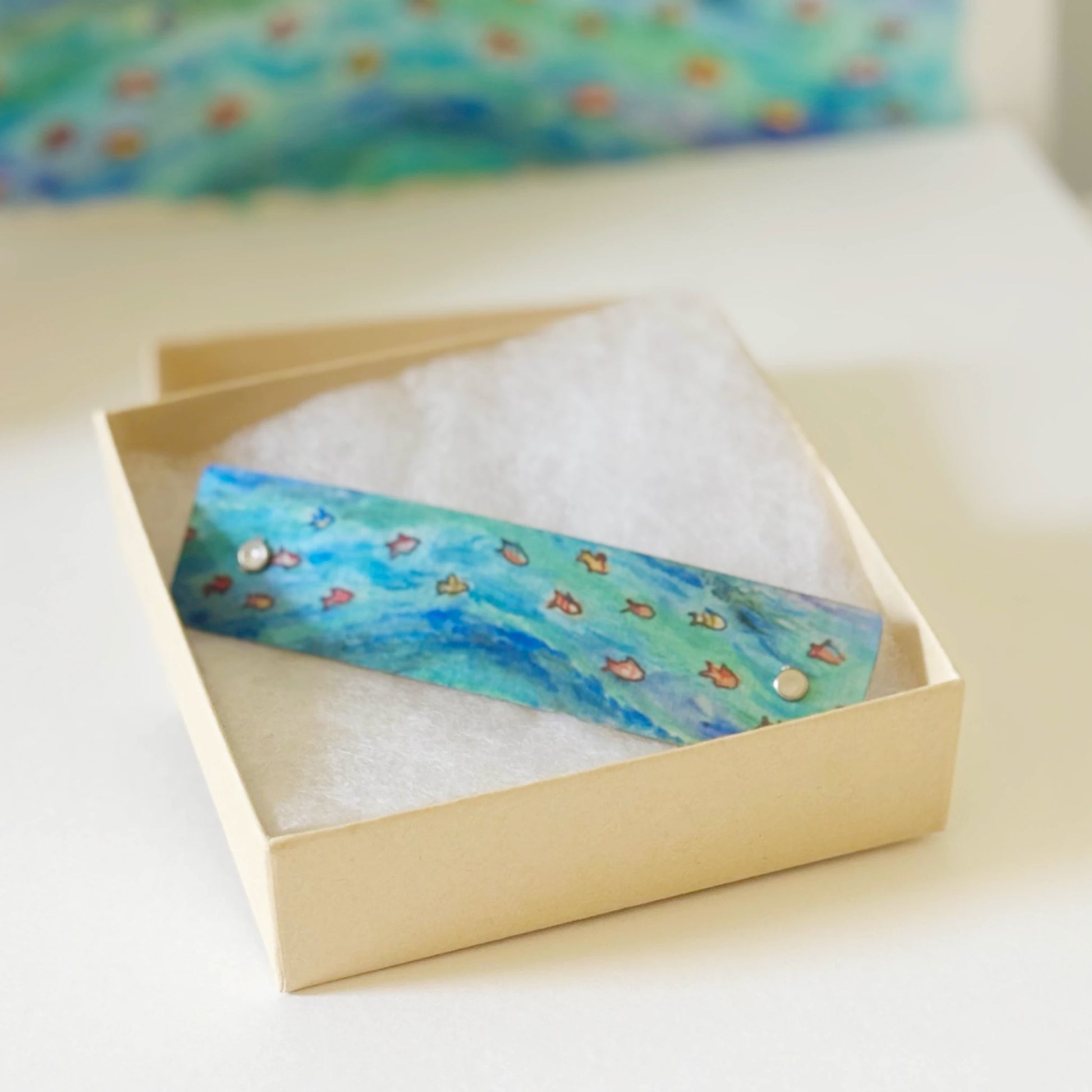 A rectangular blue and green ocean design hair barrette with small orange fish rests on white cotton inside a light beige gift box.