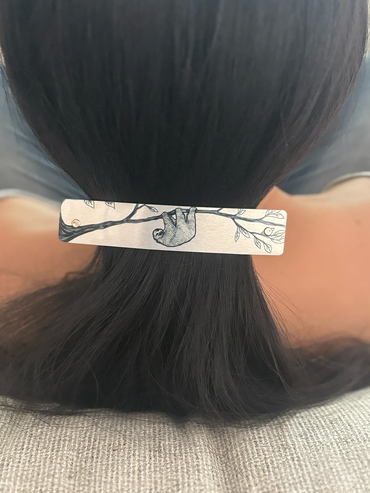 A close-up of a person's dark hair secured in a low ponytail, held with a rectangular silver metal hair clip decorated with a black line drawing of a sloth on a tree branch.