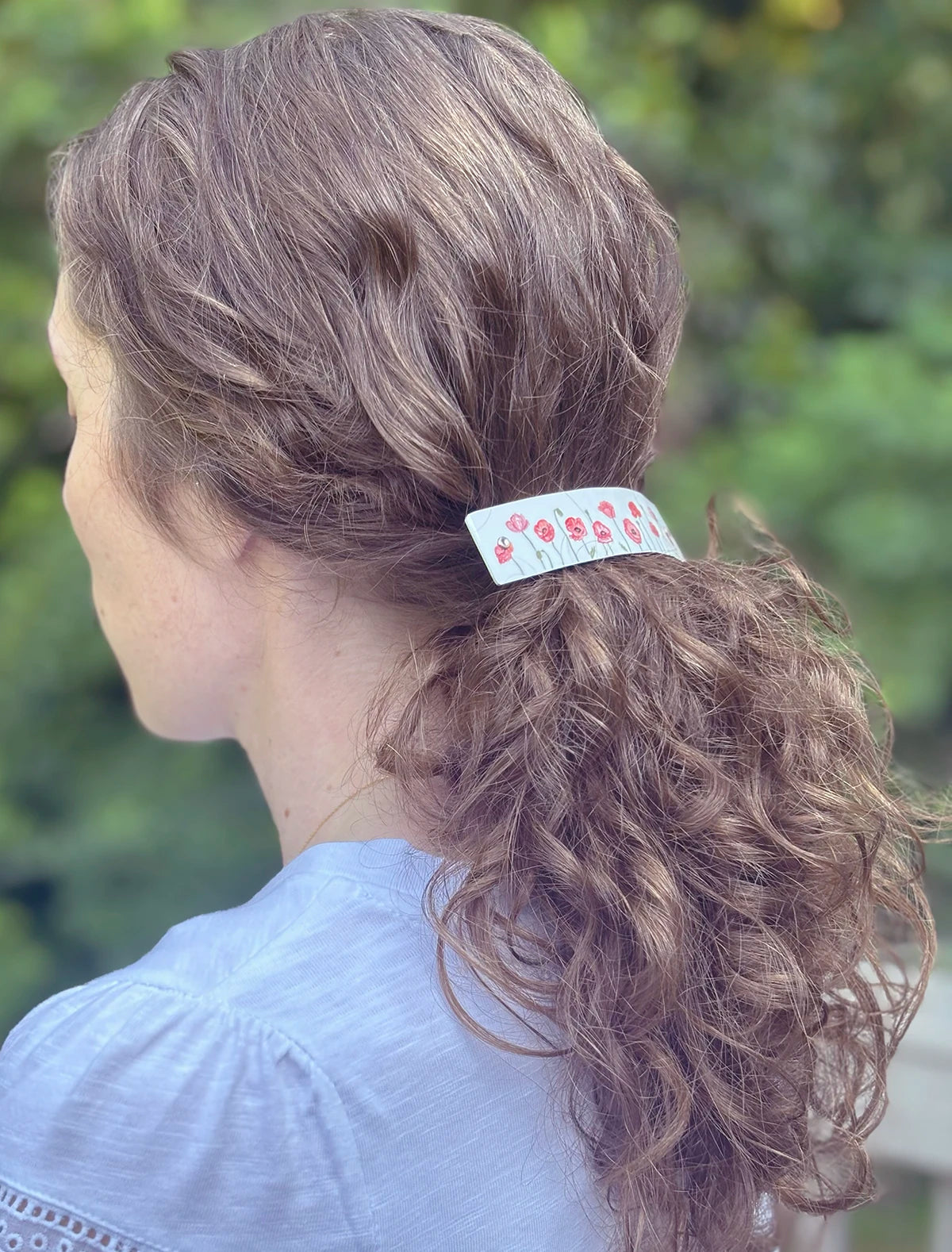 A close-up back view of a woman with curly brown hair wearing a white metal artisan hair clip adorned with red poppy illustrations, set against a blurred green outdoor background.