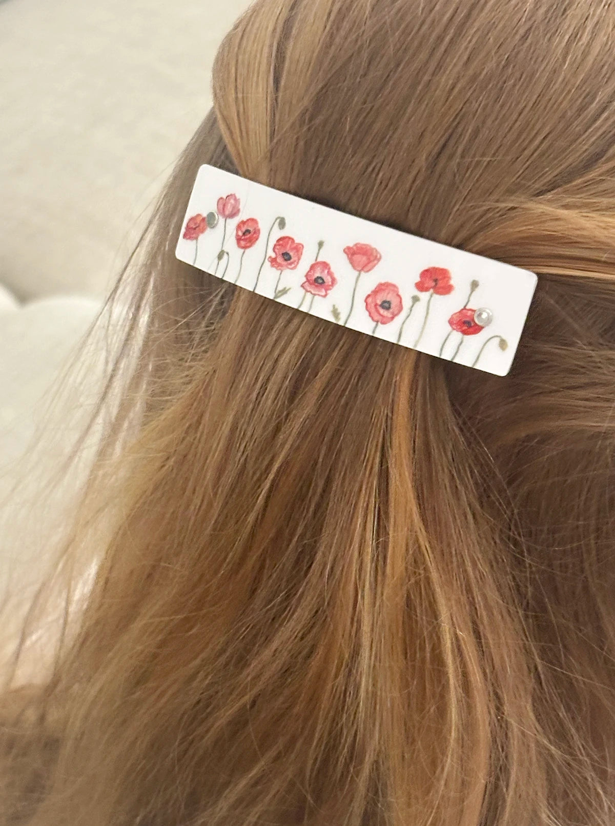 A close-up of a white rectangular hair clip decorated with red poppy flowers and green stems, worn in a half-up hairstyle on light brown hair.