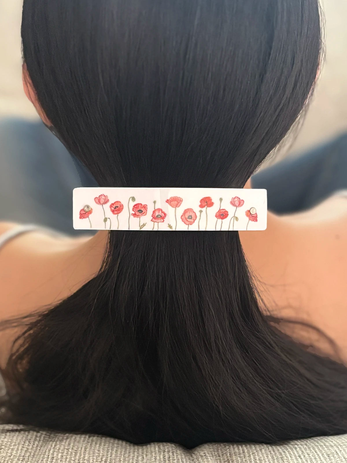 A close-up back view of a person with long black hair secured in a low ponytail, wearing a white handmade hair clip decorated with a red poppy floral watercolor pattern.