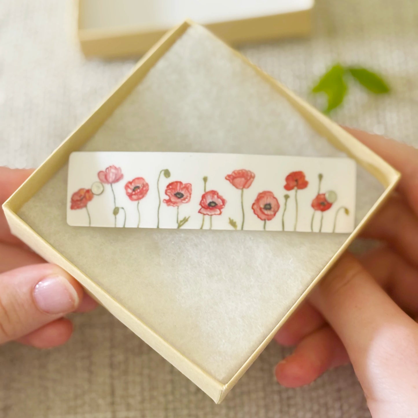 A close-up of a hand holding an open beige gift box, showing a hair clip with a row of red poppy flowers and green stems, while another hand holds the sides of the box.