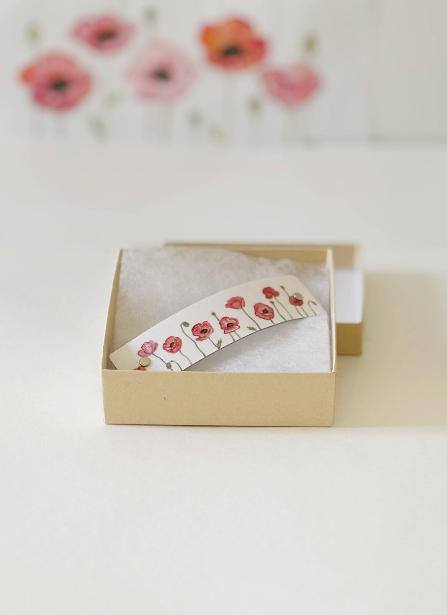 A white hair clip with a red poppy watercolor design, presented inside a small beige gift box with cotton, on a white surface with the original art in the blurred background.