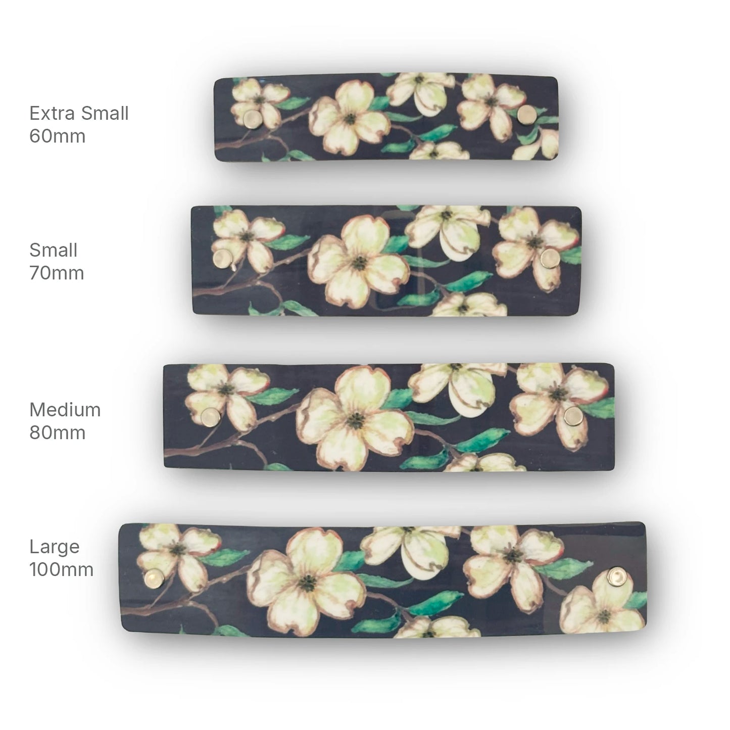 Four rectangular floral French barrettes are  shown in different sizes labeled Extra Small 60mm, Small 70mm, Medium 80mm, and Large 100mm. The hair clips have a dark charcoal background with cream-colored dogwood flowers and green leaves.