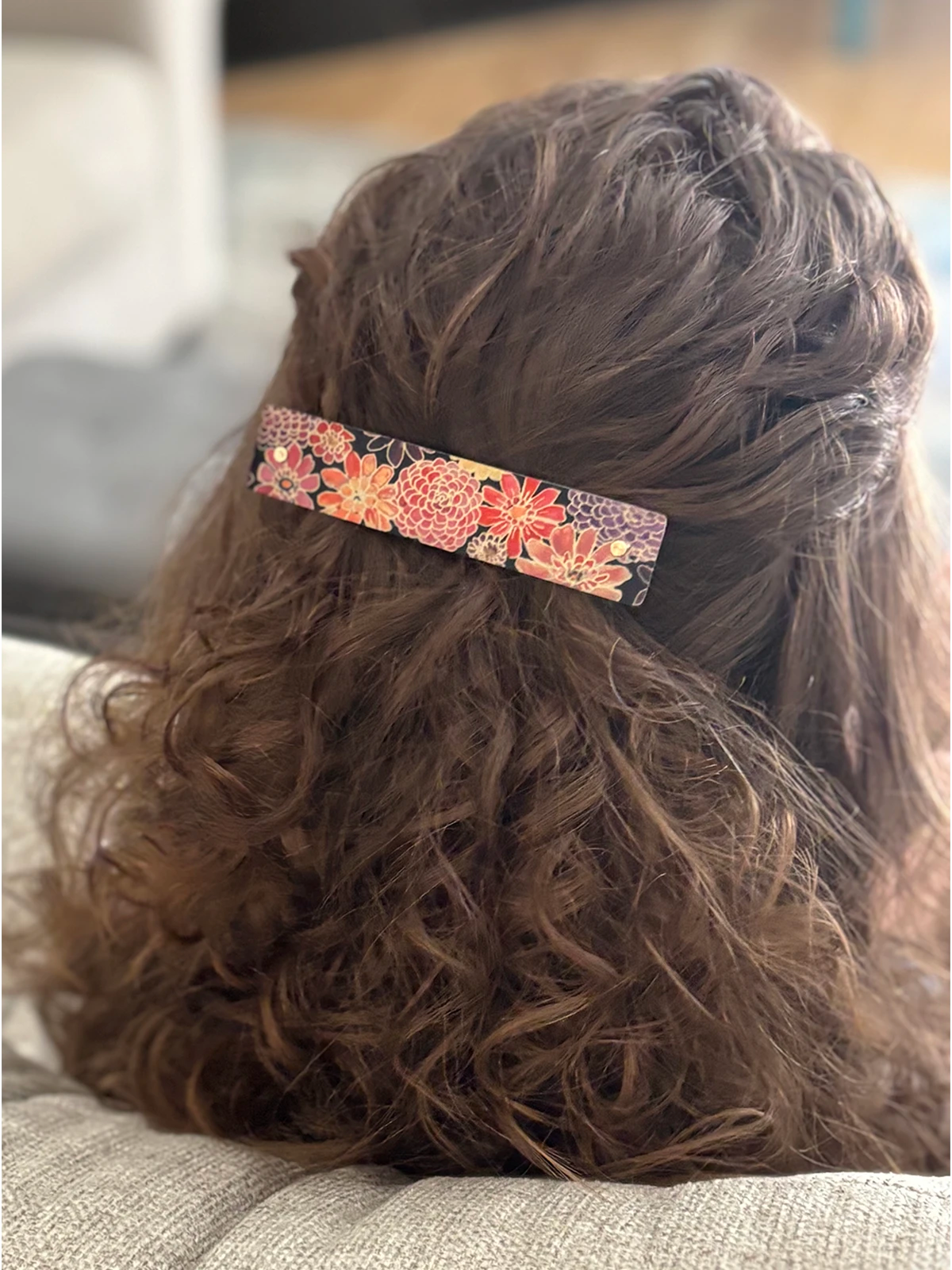 A close-up back view of a person with long, wavy brown hair wearing a rectangular hair clip adorned with a colorful floral pattern in orange, pink, and purple.