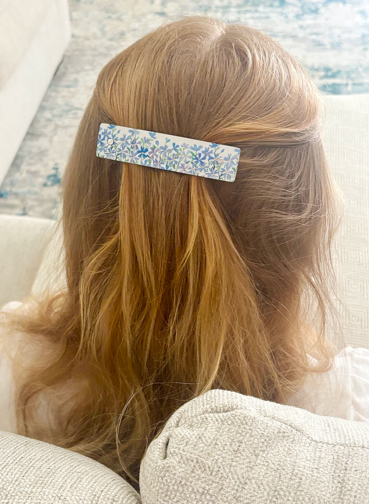 A close-up back view of a person with long blonde hair wearing a rectangular hair clip with a blue and purple wildflower pattern, seated on a light-colored couch.
