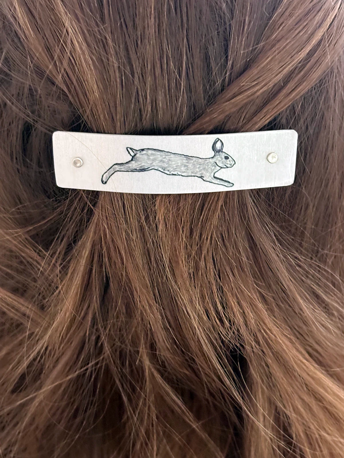 A close-up of a silver-toned metal hair clip featuring a realistic original rabbit design, shown secured in brown hair.