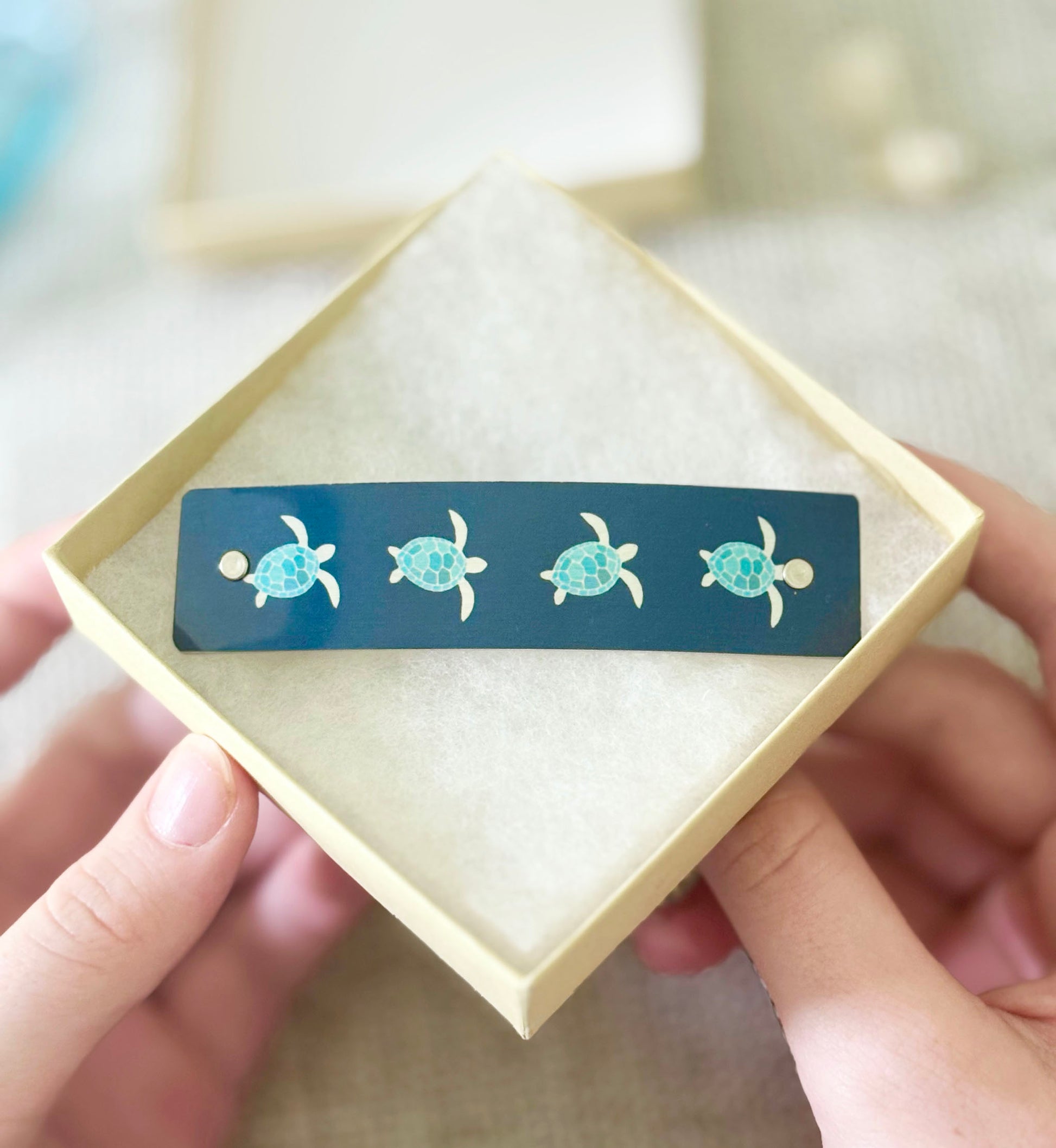 A close-up of hands holding a square cream-colored gift box containing a long, dark teal bar decorated with four small turquoise and silver sea turtle designs.