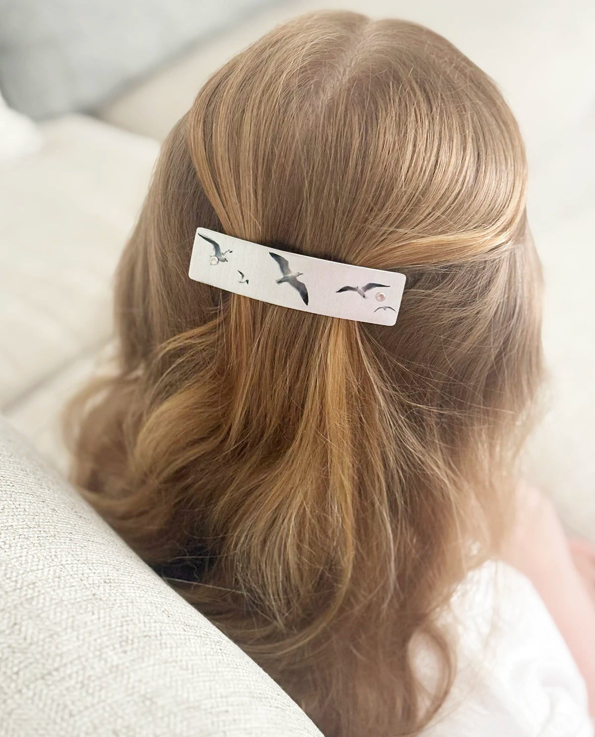 A close-up lifestyle shot of a woman's blonde hair secured with a silver rectangular hair clip decorated with seagulls, resting on a light-colored sofa.