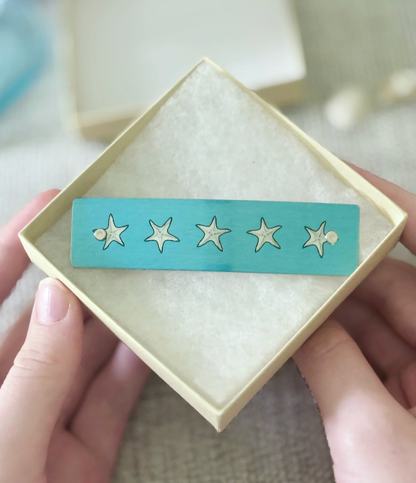 A person's hands holding a square cream-colored jewelry box, displaying a turquoise blue bar with five gold star designs.