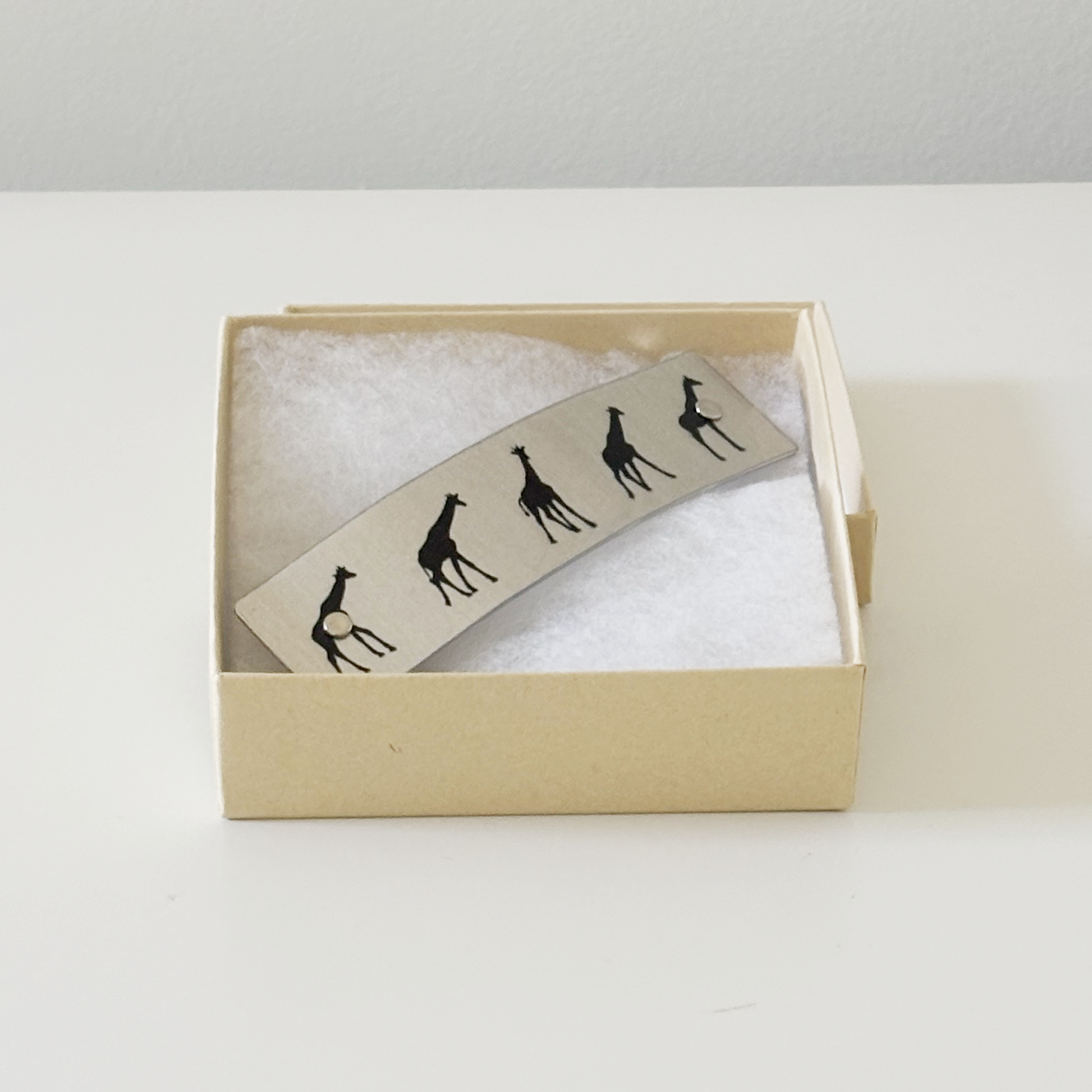 A silver-toned metal hair clip featuring black silhouettes of five giraffes, presented inside a light beige gift box with white cotton on a white surface.
