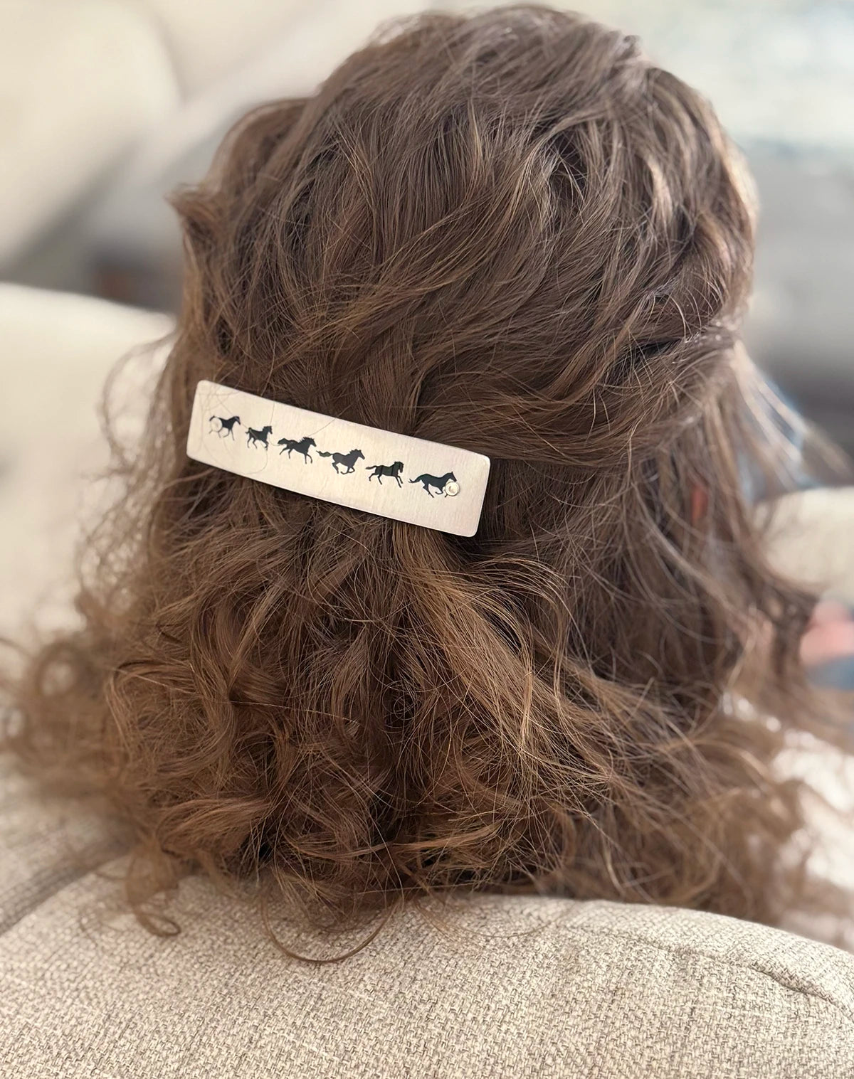 A close-up of a person's brown curly hair from behind, featuring a silver rectangular hair clip adorned with black horse silhouettes.