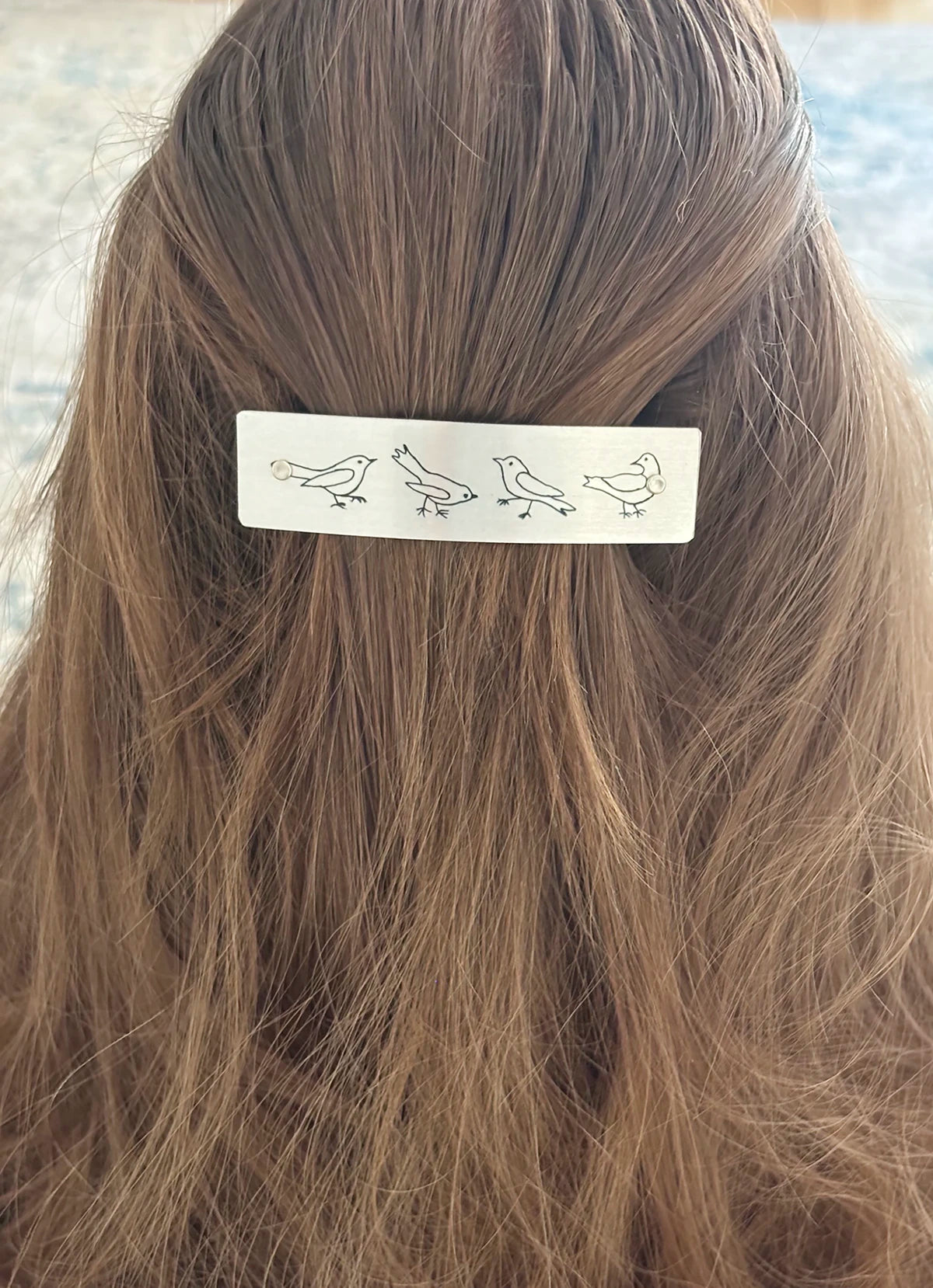 A close-up of a person's brown hair secured with a silver rectangular hair clip decorated with simple black bird illustrations.