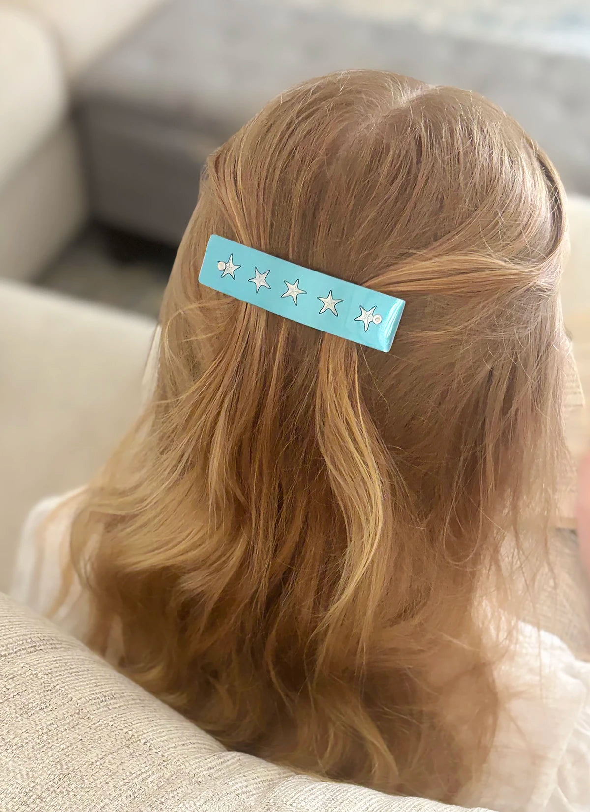 A light blue hair clip with five silver starfish designs, holding back a blonde girl's hair, shown from behind.