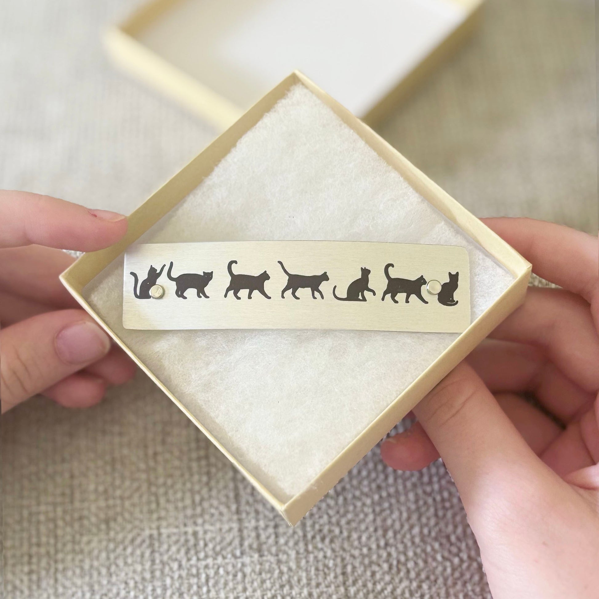 A hair clip featuring black cat silhouettes, held in a square cream gift box with white tissue paper, being held by two hands over a textured beige fabric background.