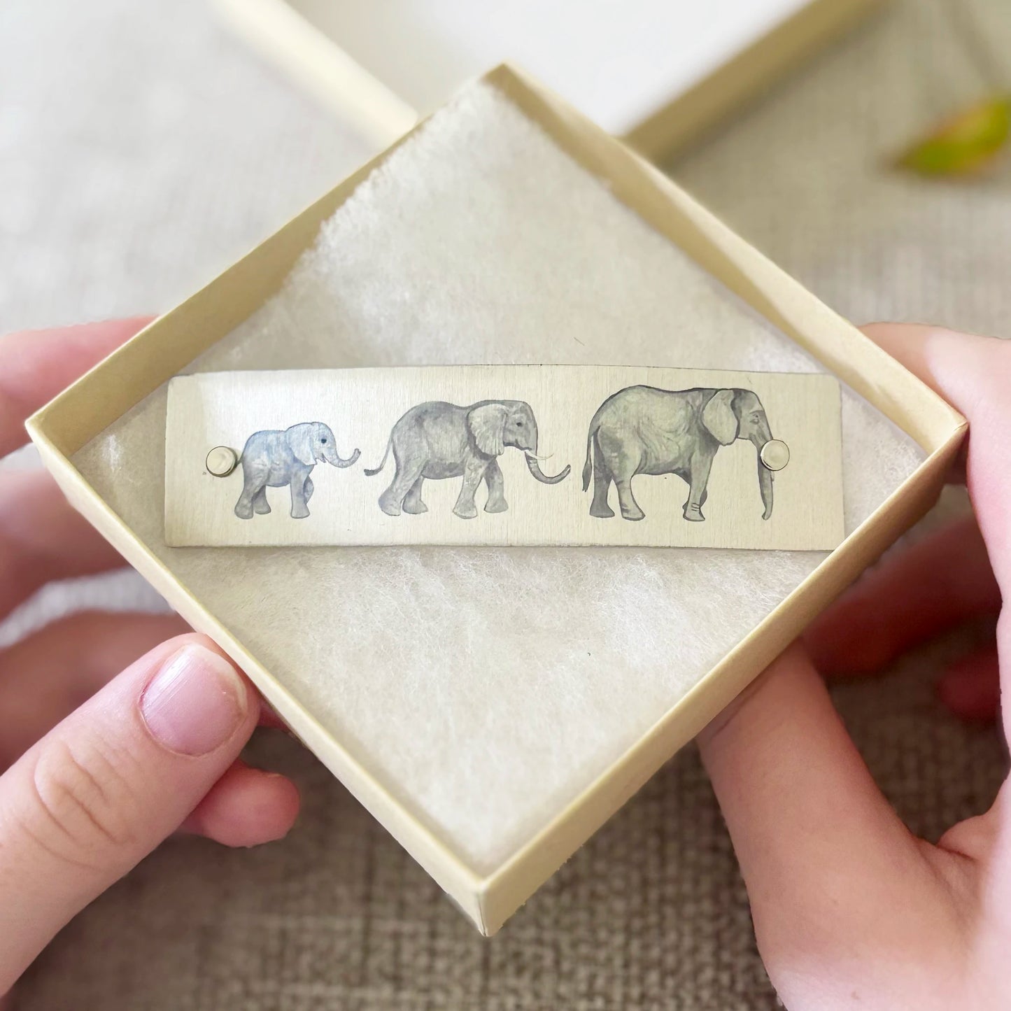 A close-up of hands holding an open beige gift box containing a metal French barrette with illustrated elephants.