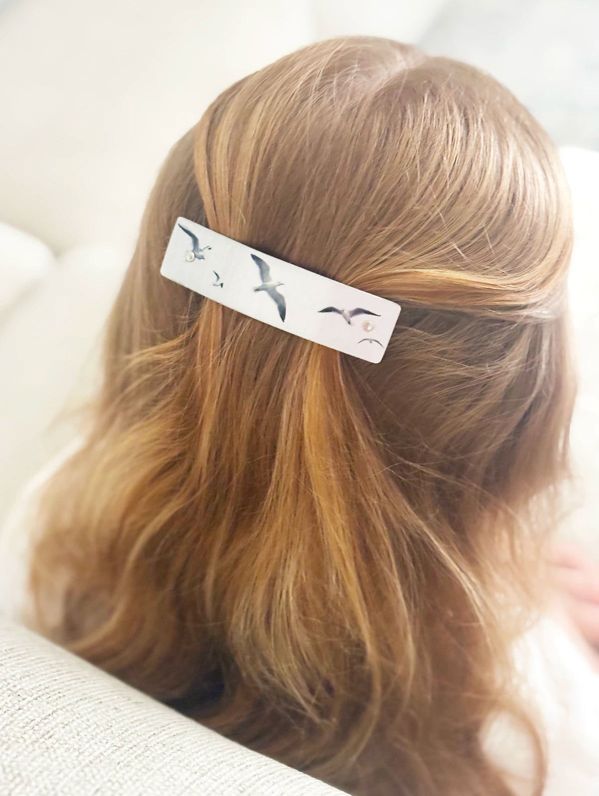 A close-up lifestyle shot of a person with light brown hair wearing a silver hair clip decorated with seagull illustrations.